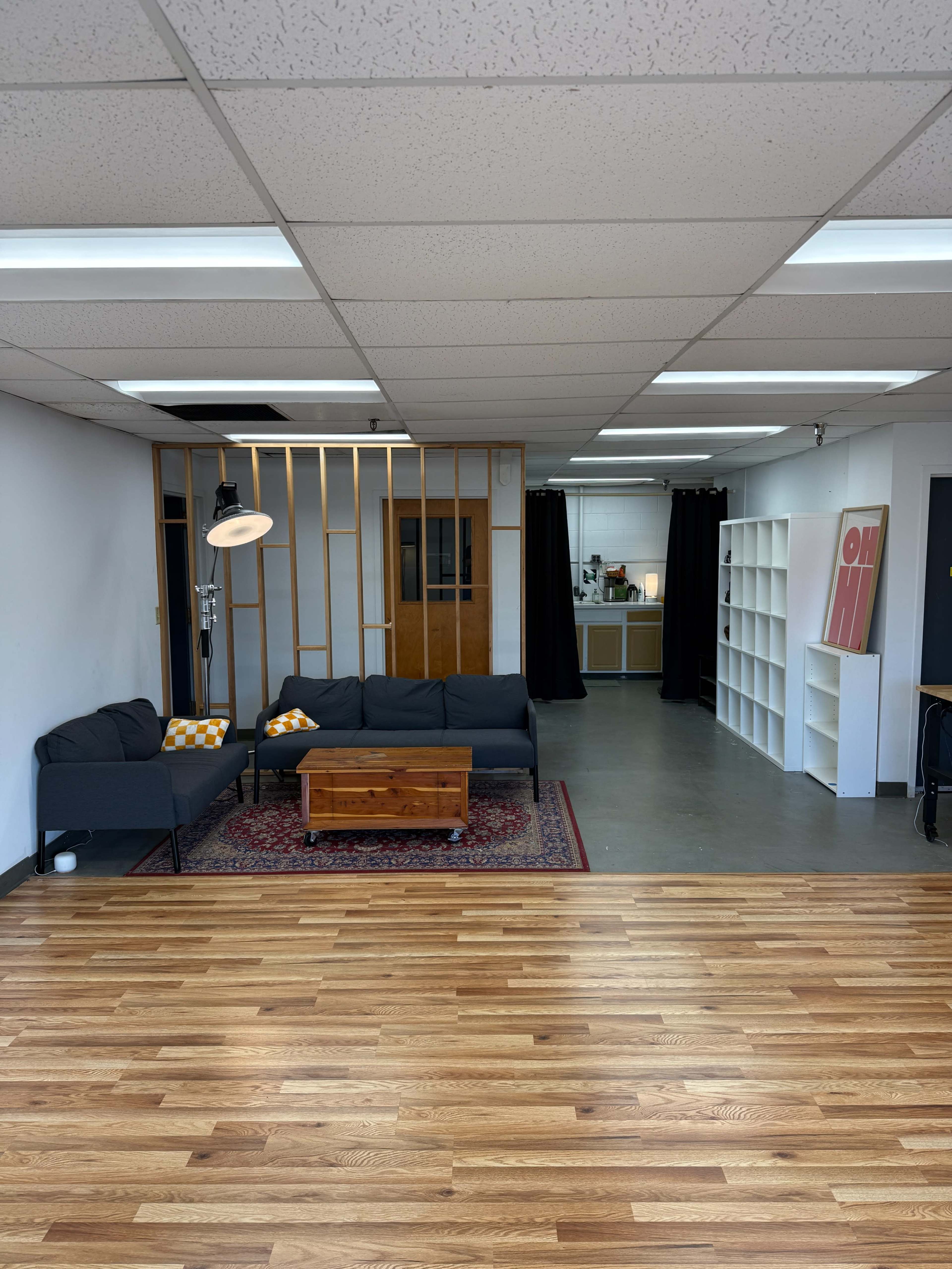The image shows a spacious room featuring a seating area with a sofa and coffee table, wood flooring, and an open kitchen area in the background.