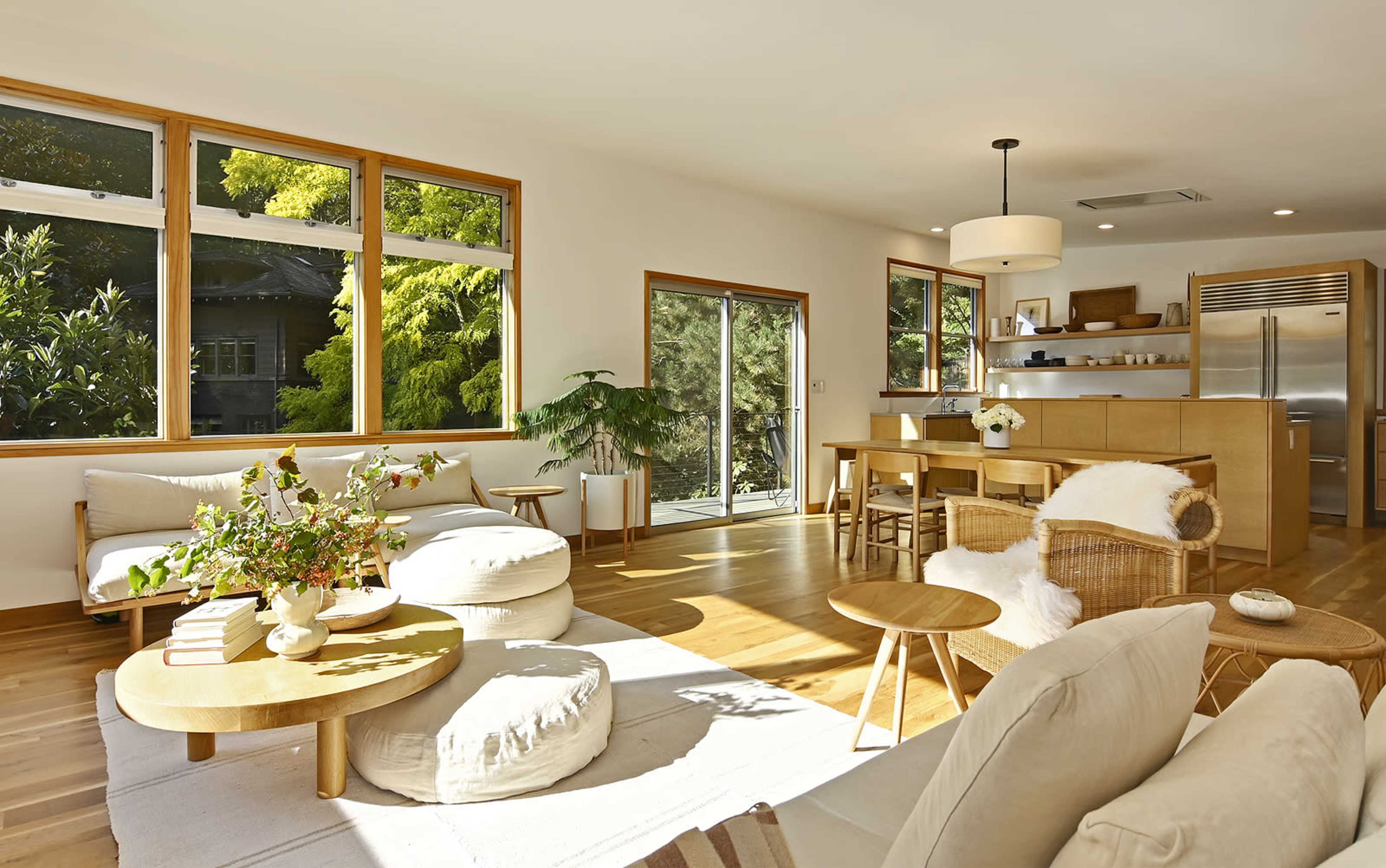 A bright living room features large windows, wooden furniture, and a dining area connected to an open kitchen.