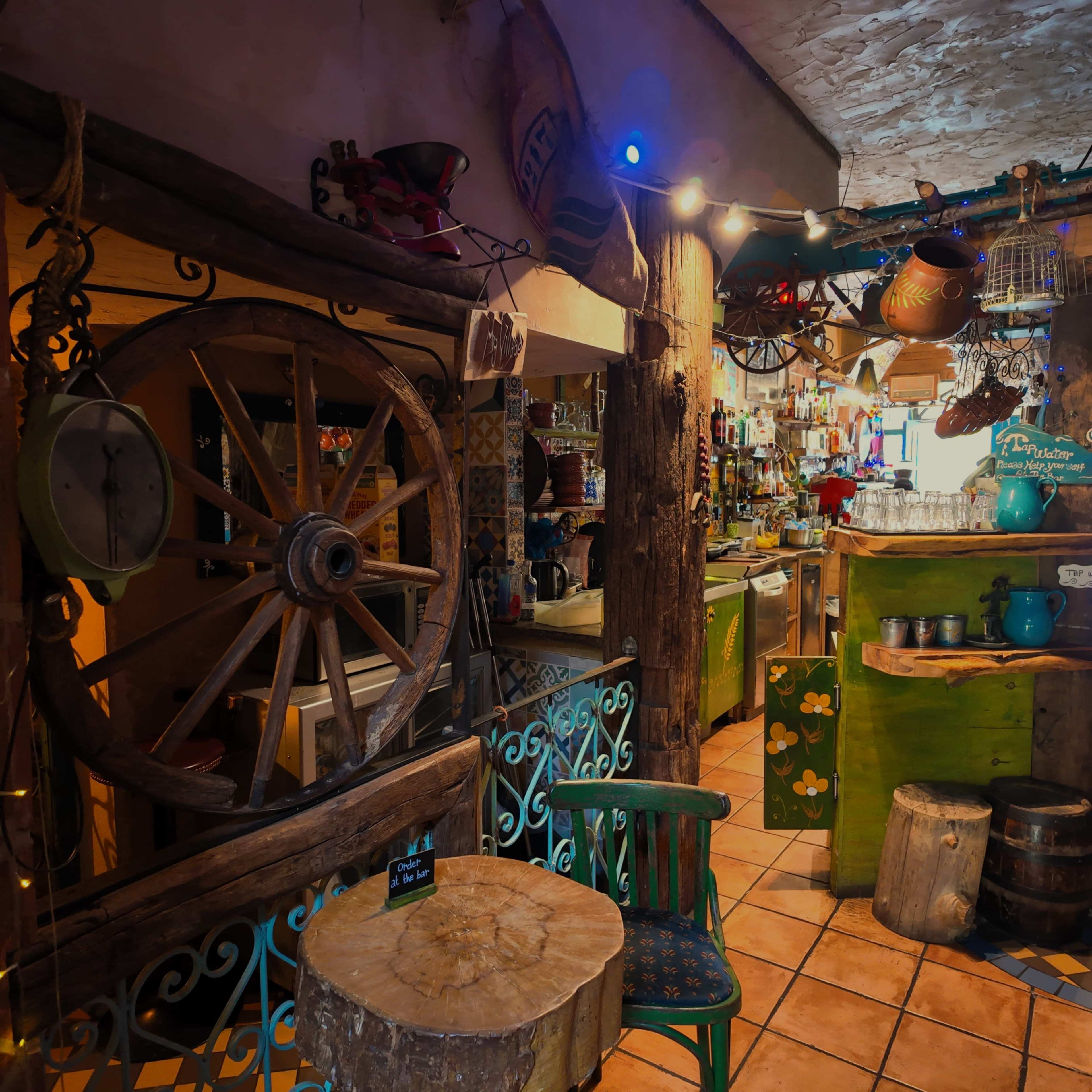 The image depicts a rustic interior of a cozy café or bar featuring wooden décor, a large wagon wheel, and various kitchen items arranged on shelves.