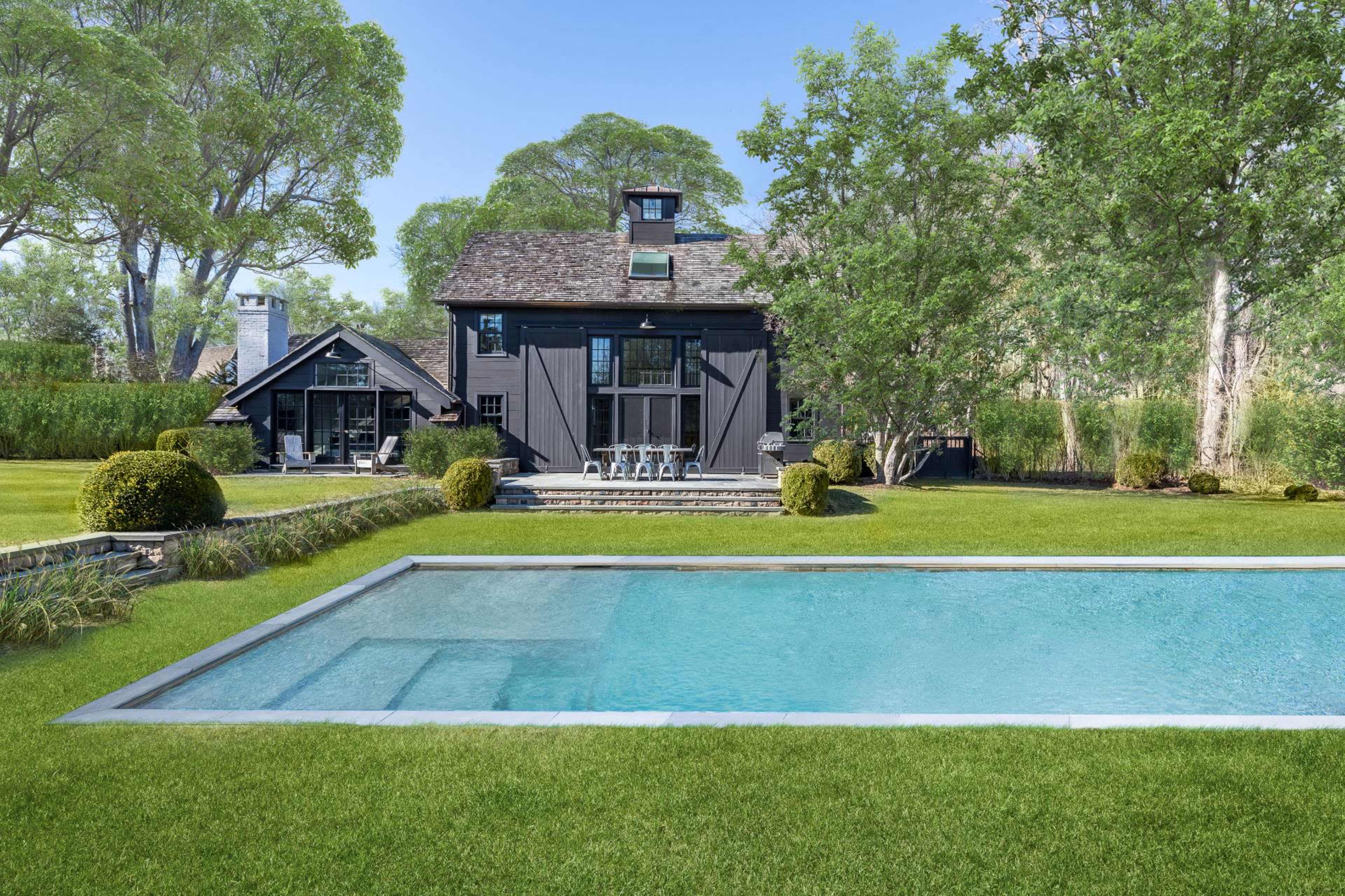A modern black house with large windows is situated beside a rectangular swimming pool in a well-maintained lawn surrounded by trees.