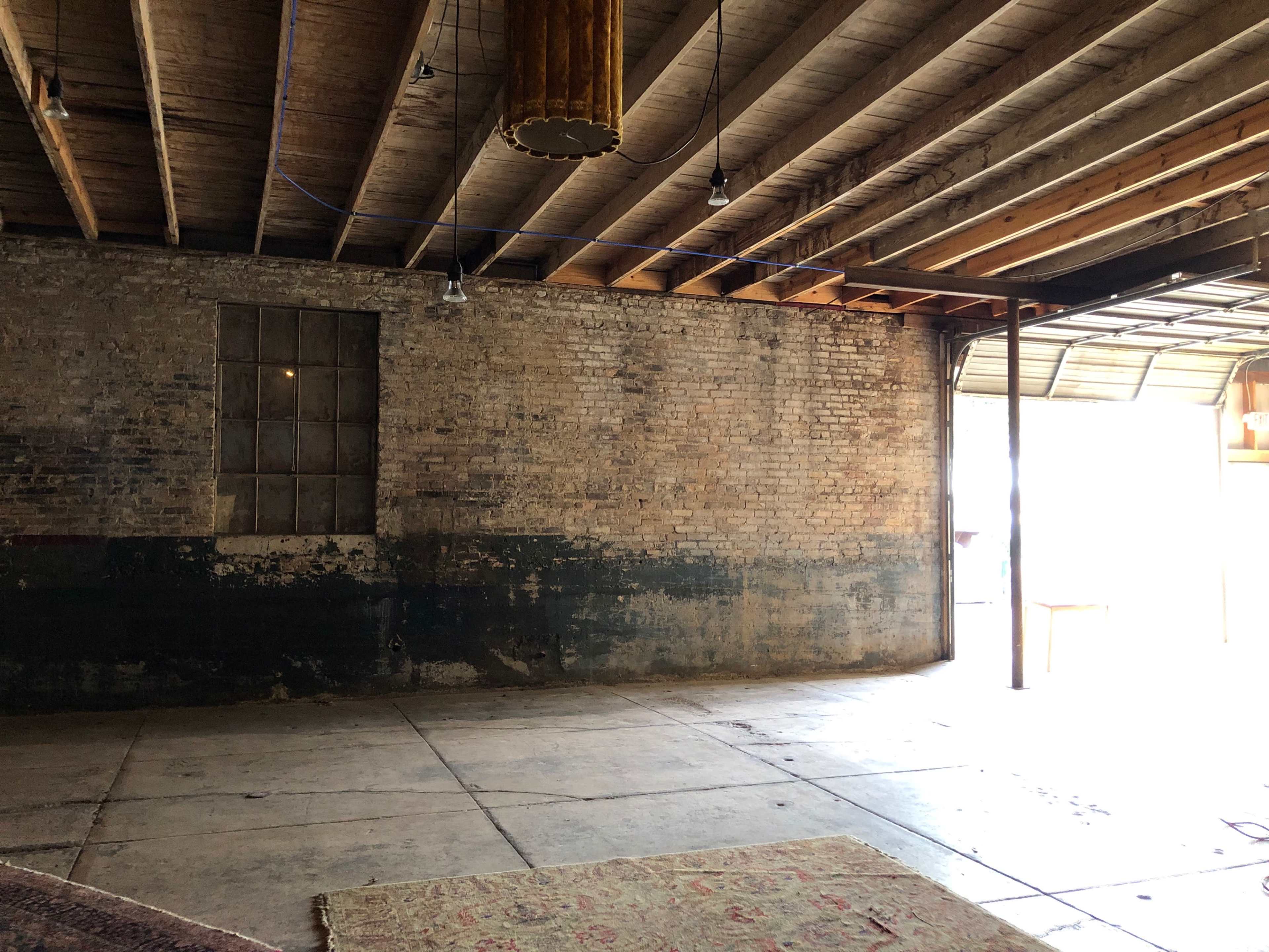 The image shows an empty, industrial-style room with exposed brick walls, a concrete floor, and a large opening leading outside.