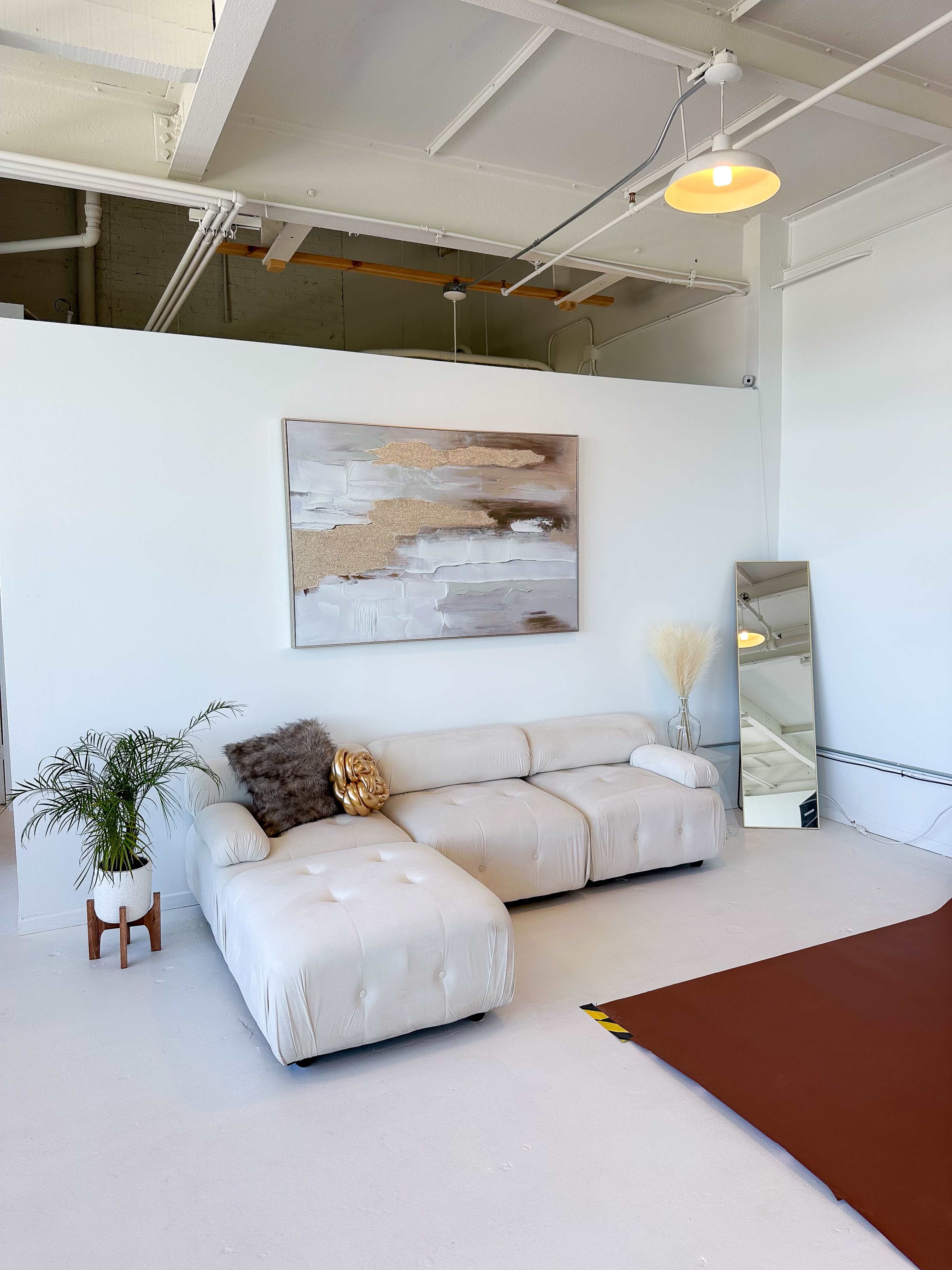 A modern living space features a light-colored sofa, a decorative painting on the wall, a mirror, and a small plant on a stand.