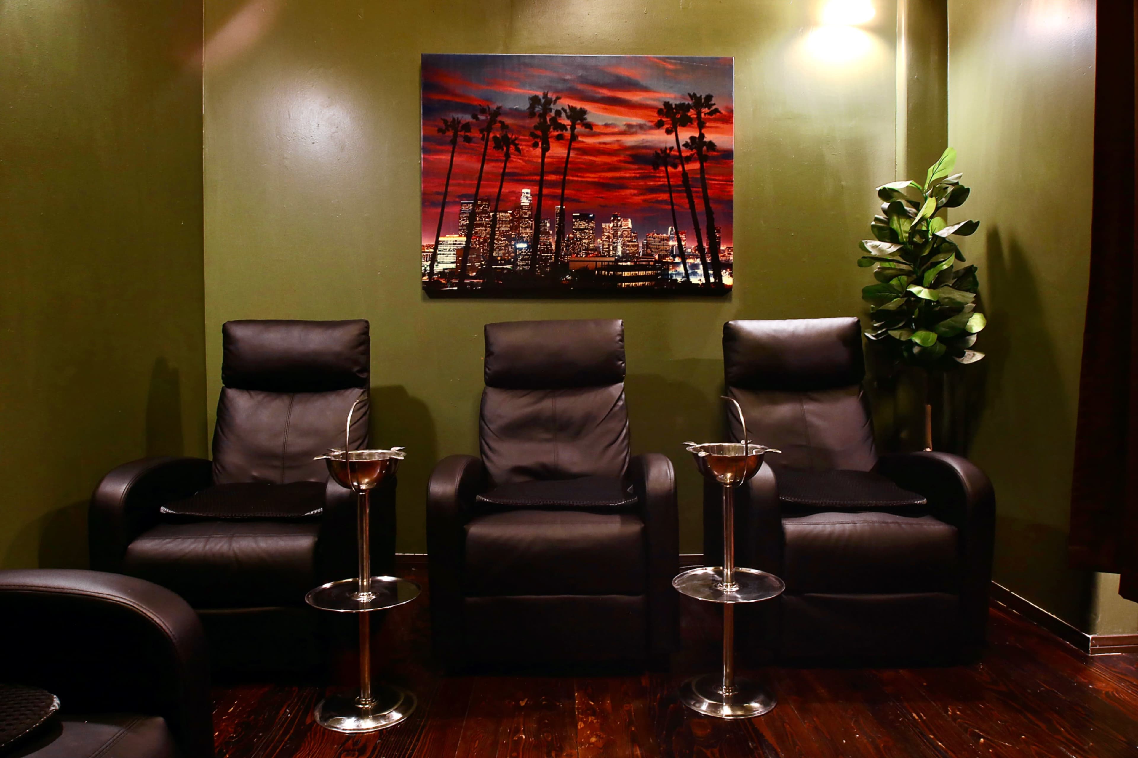 Three black leather chairs with side tables, positioned under a framed sunset cityscape with palm trees on a green wall.