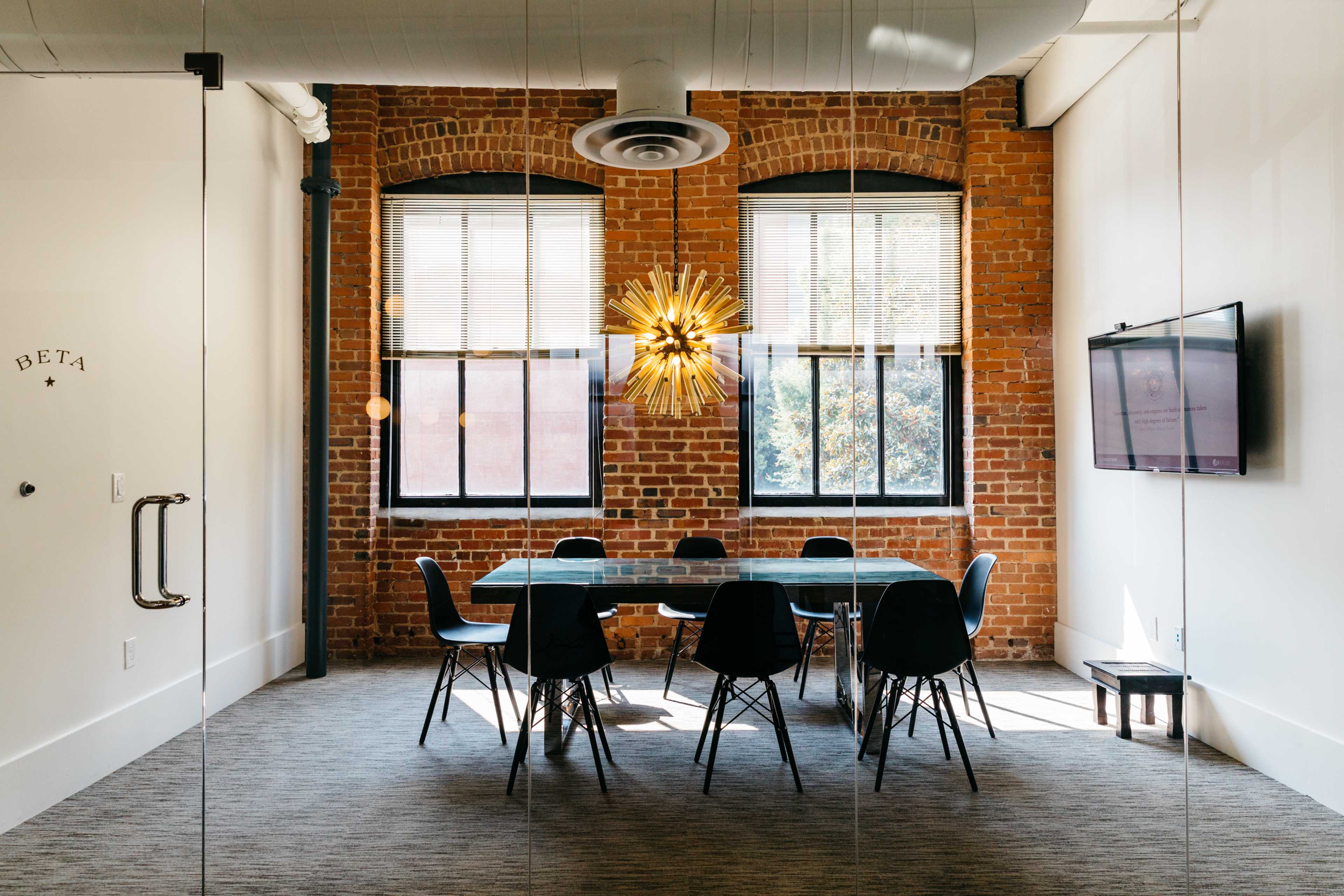 A modern conference room features a large table surrounded by black chairs, with exposed brick walls and large windows allowing natural light to enter.