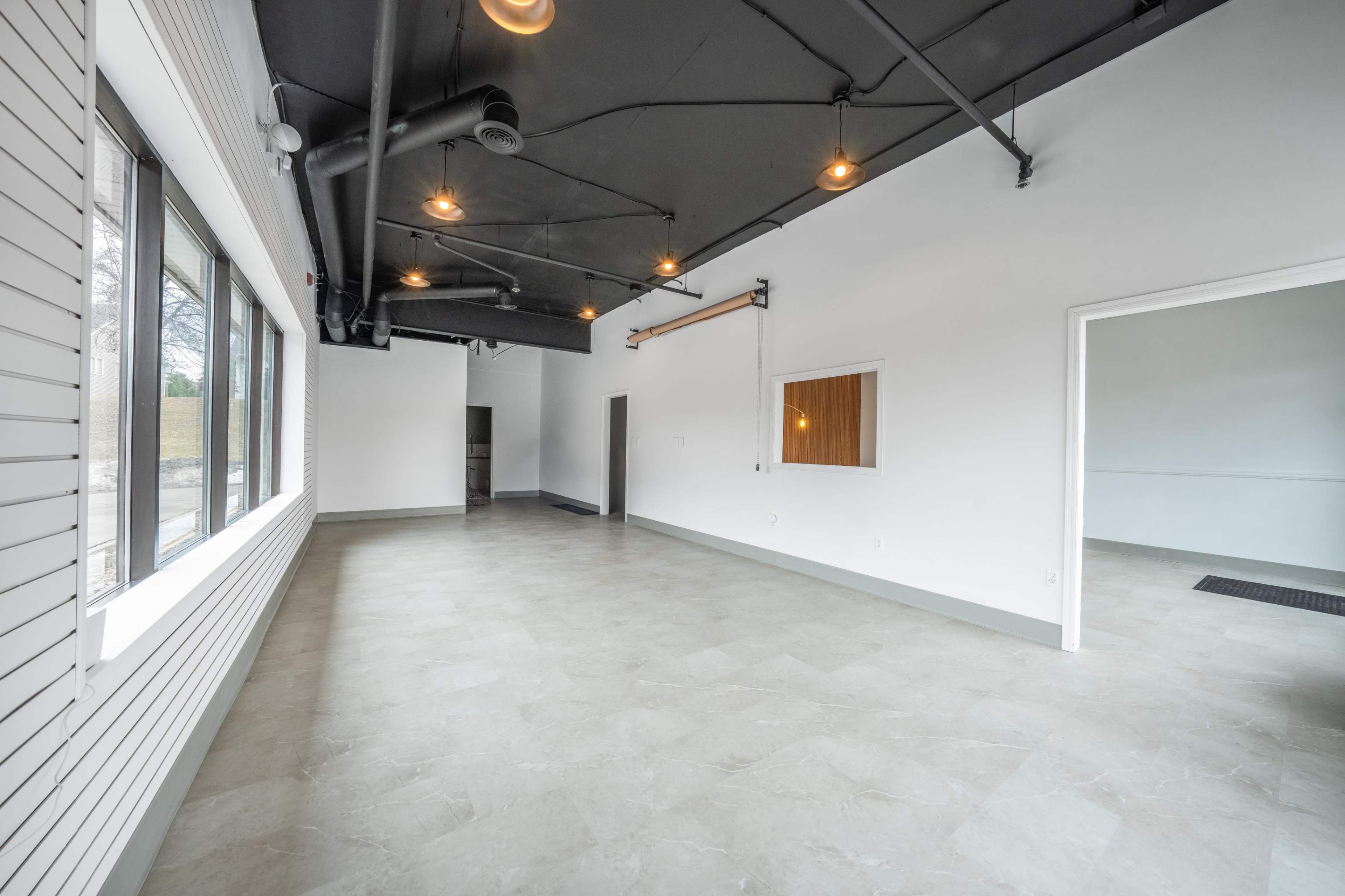 The image shows an empty commercial space with tiled flooring, white walls, and large windows allowing natural light to enter.