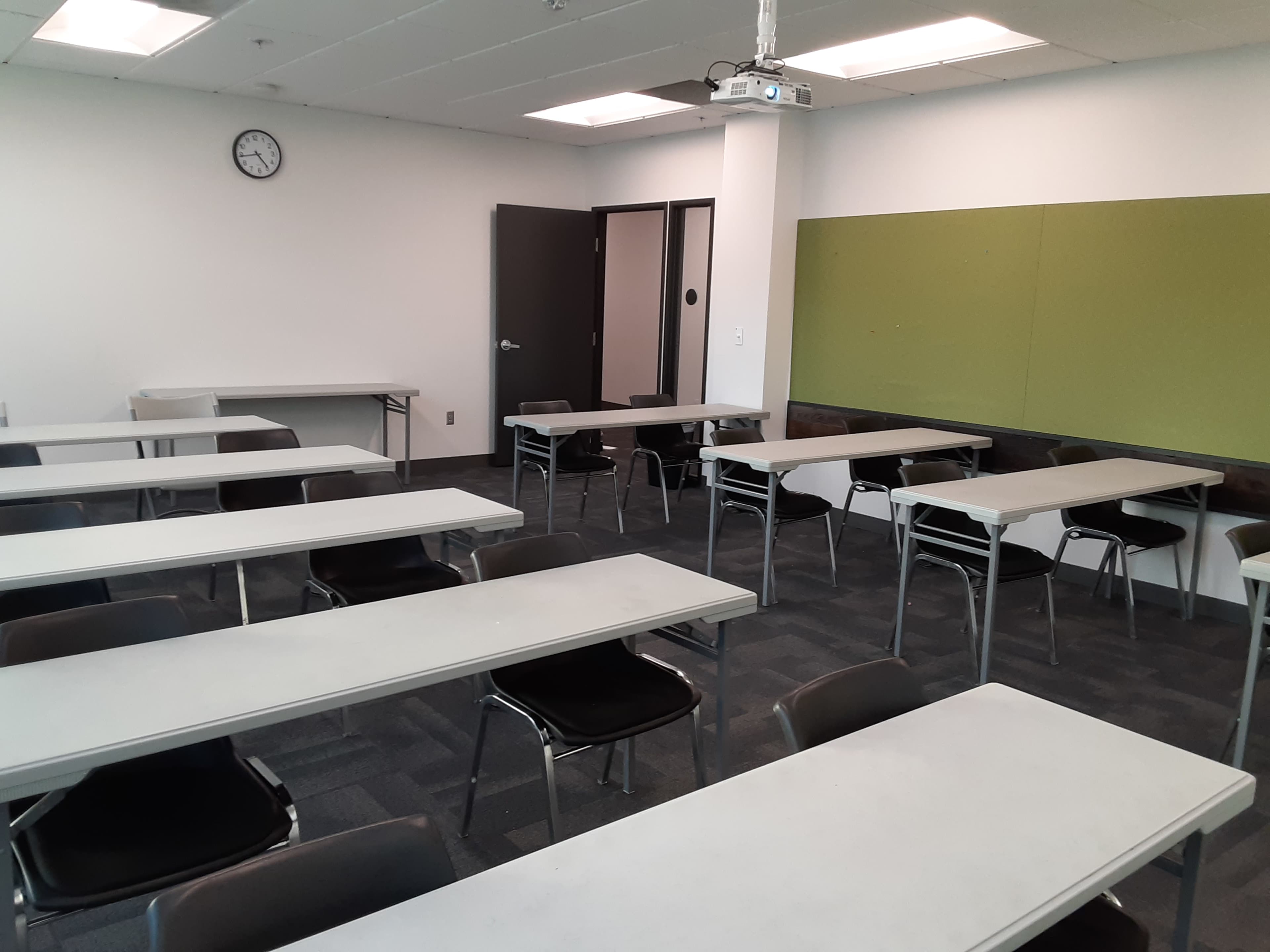 A classroom with rows of tables and chairs, a clock on the wall, and a projector mounted on the ceiling.