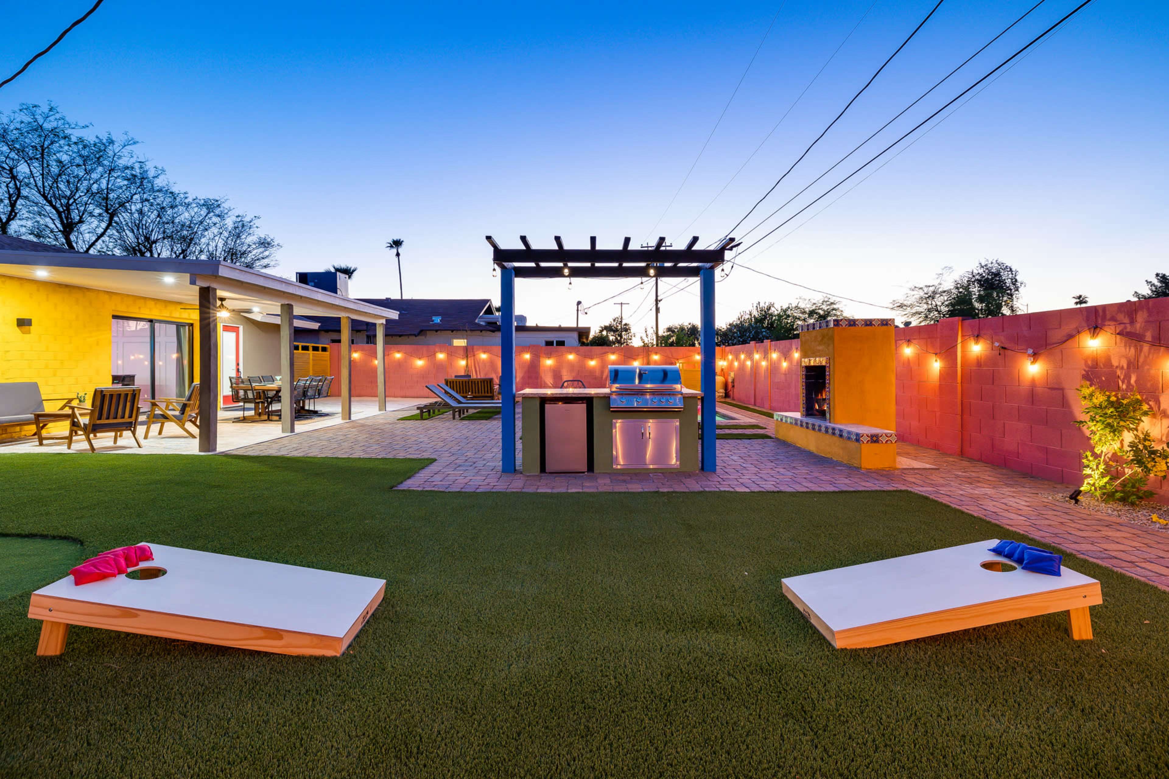 A backyard with a grill station under a pergola, two cornhole boards, and string lights illuminating the area at dusk.