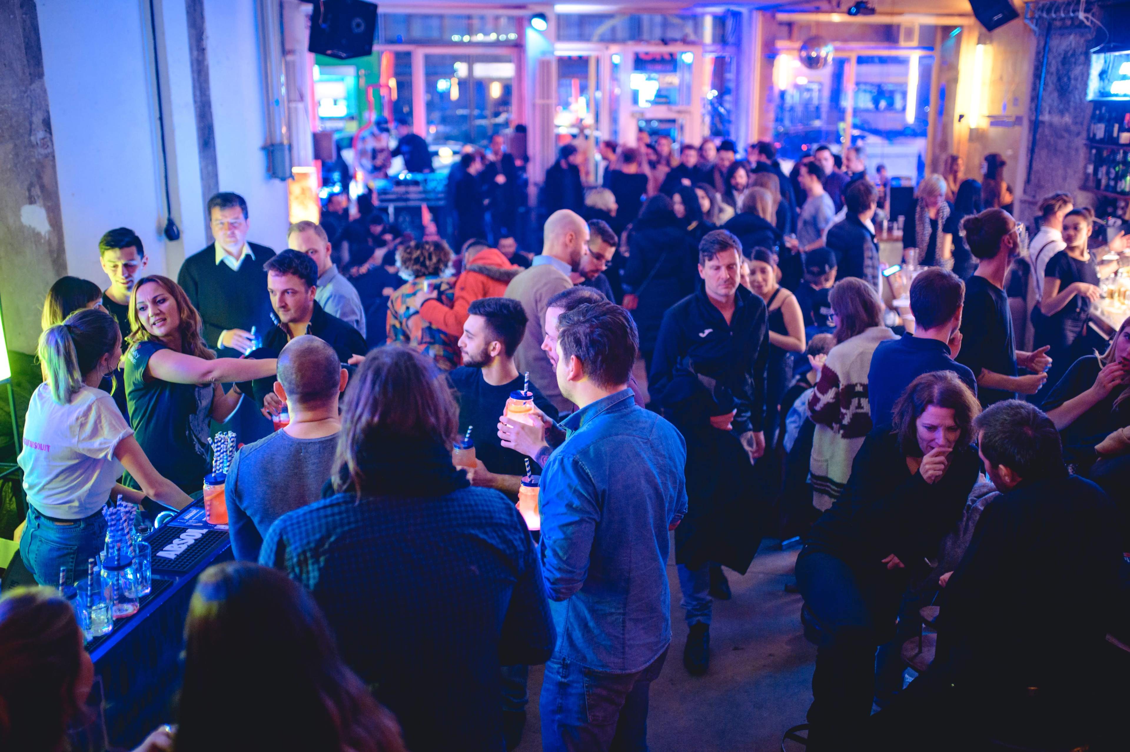 A lively crowd gathers inside a bustling bar, with people engaged in conversation and serving drinks amidst colorful lighting.