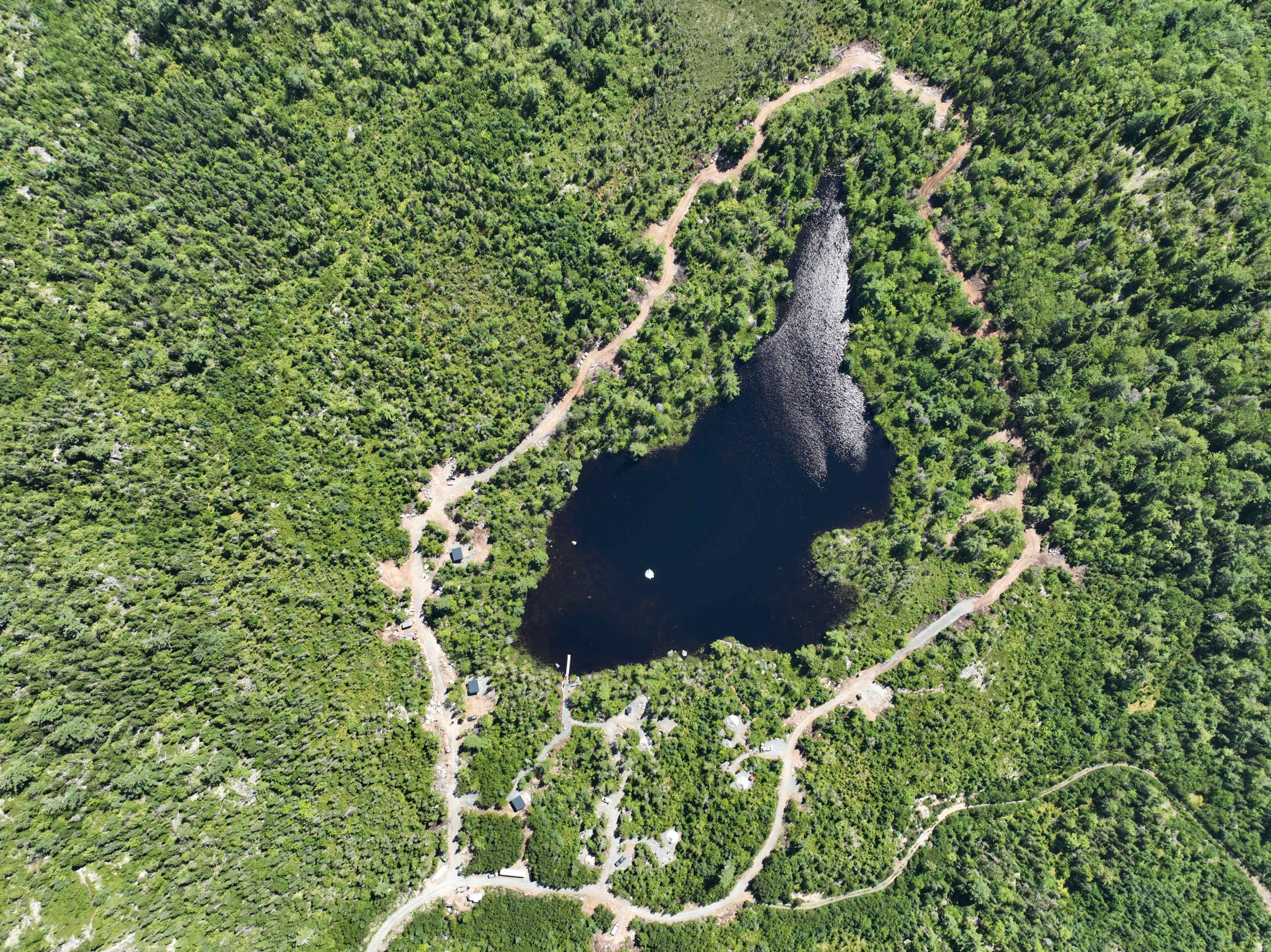 An aerial view shows a dark lake surrounded by dense greenery and winding paths in a forested area.