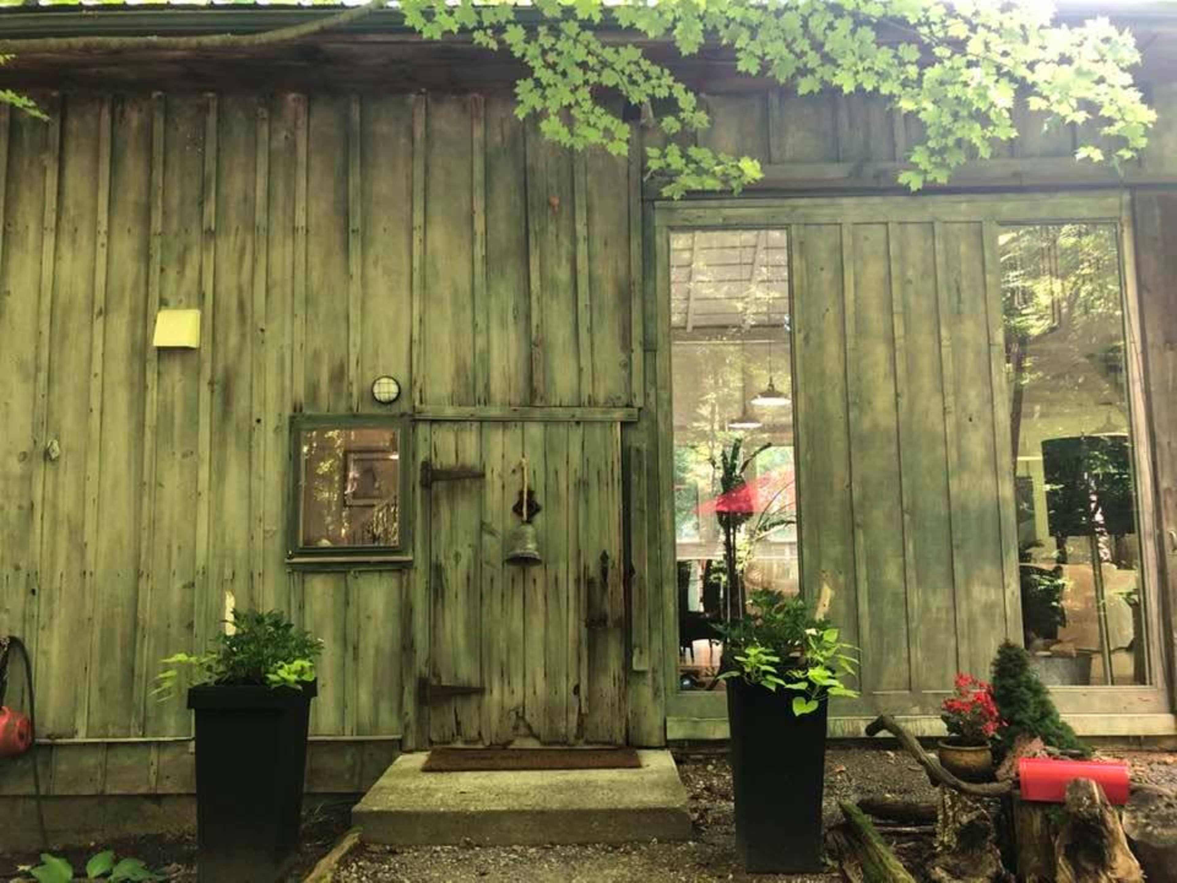 A wooden cabin with a green, weathered exterior, featuring a small door, a clock, and two planters on either side of the entrance.