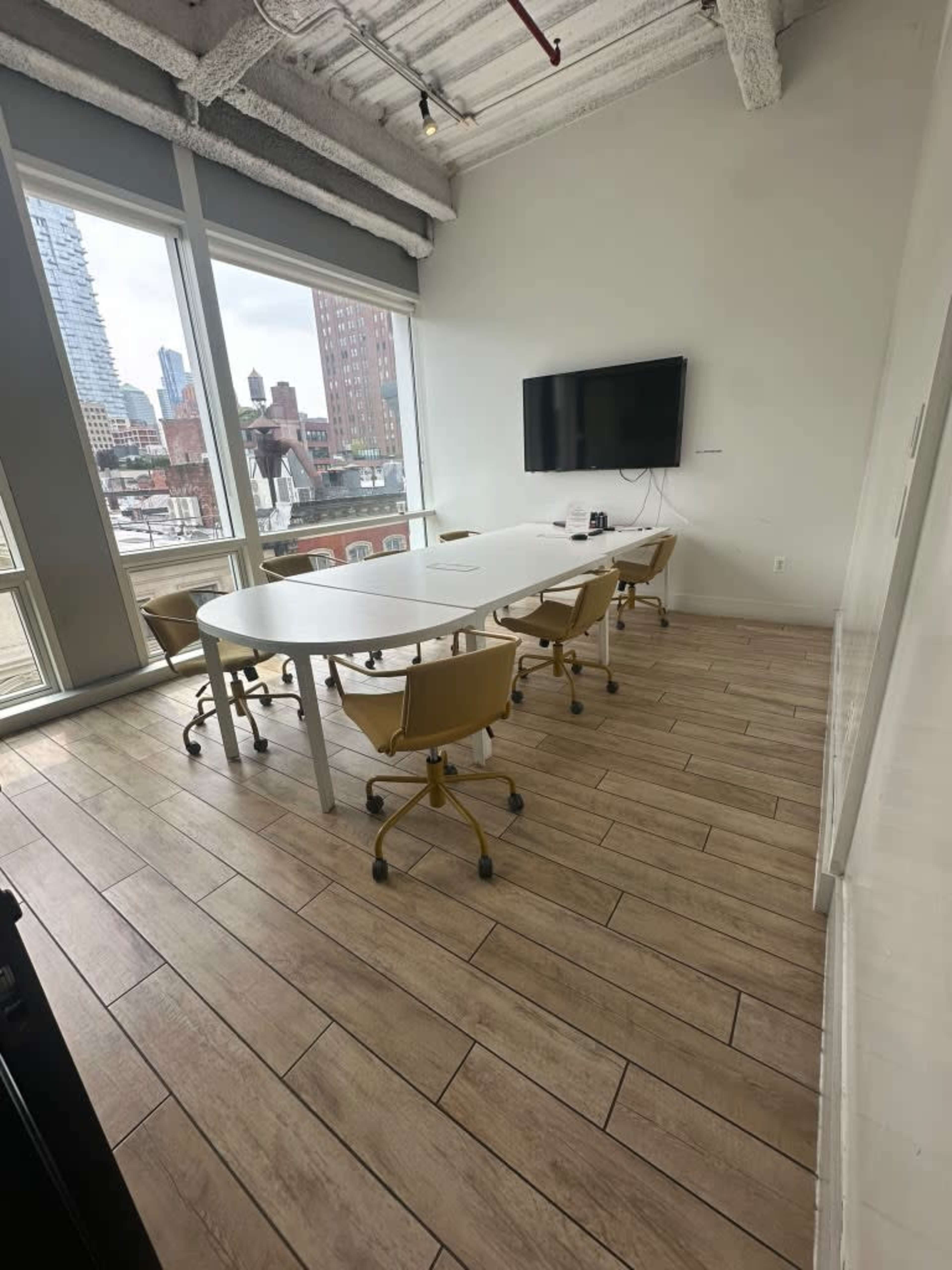 The image shows a modern conference room with a large white table, several yellow swivel chairs, and a wall-mounted television, featuring large windows that overlook a cityscape.
