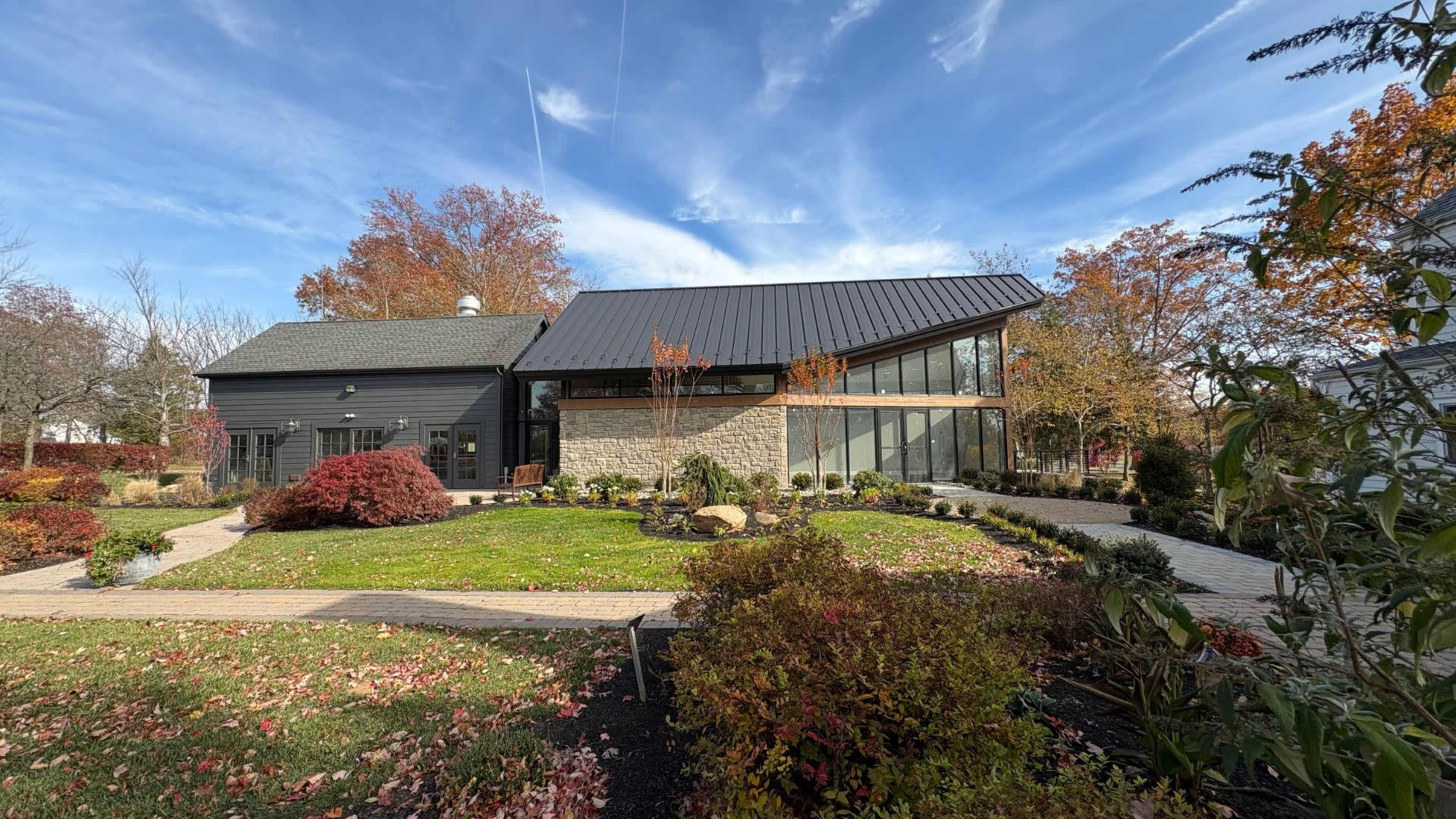 A modern house with a sleek metal roof and large windows is surrounded by landscaped gardens featuring colorful autumn foliage.