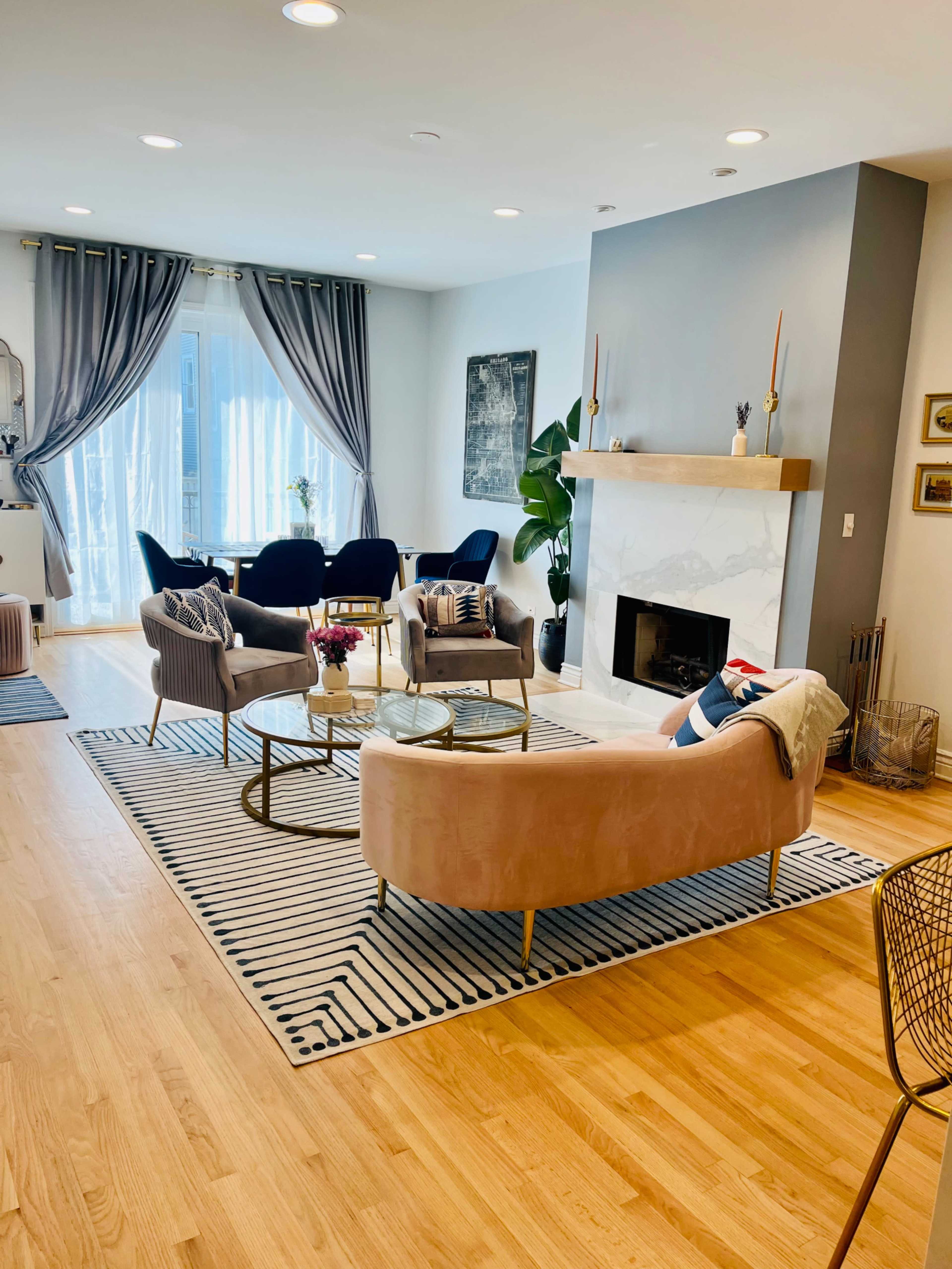 A modern living room features a light wood floor, a marble fireplace, and seating arrangements with two pink sofas, three armchairs, and a coffee table on a striped rug.