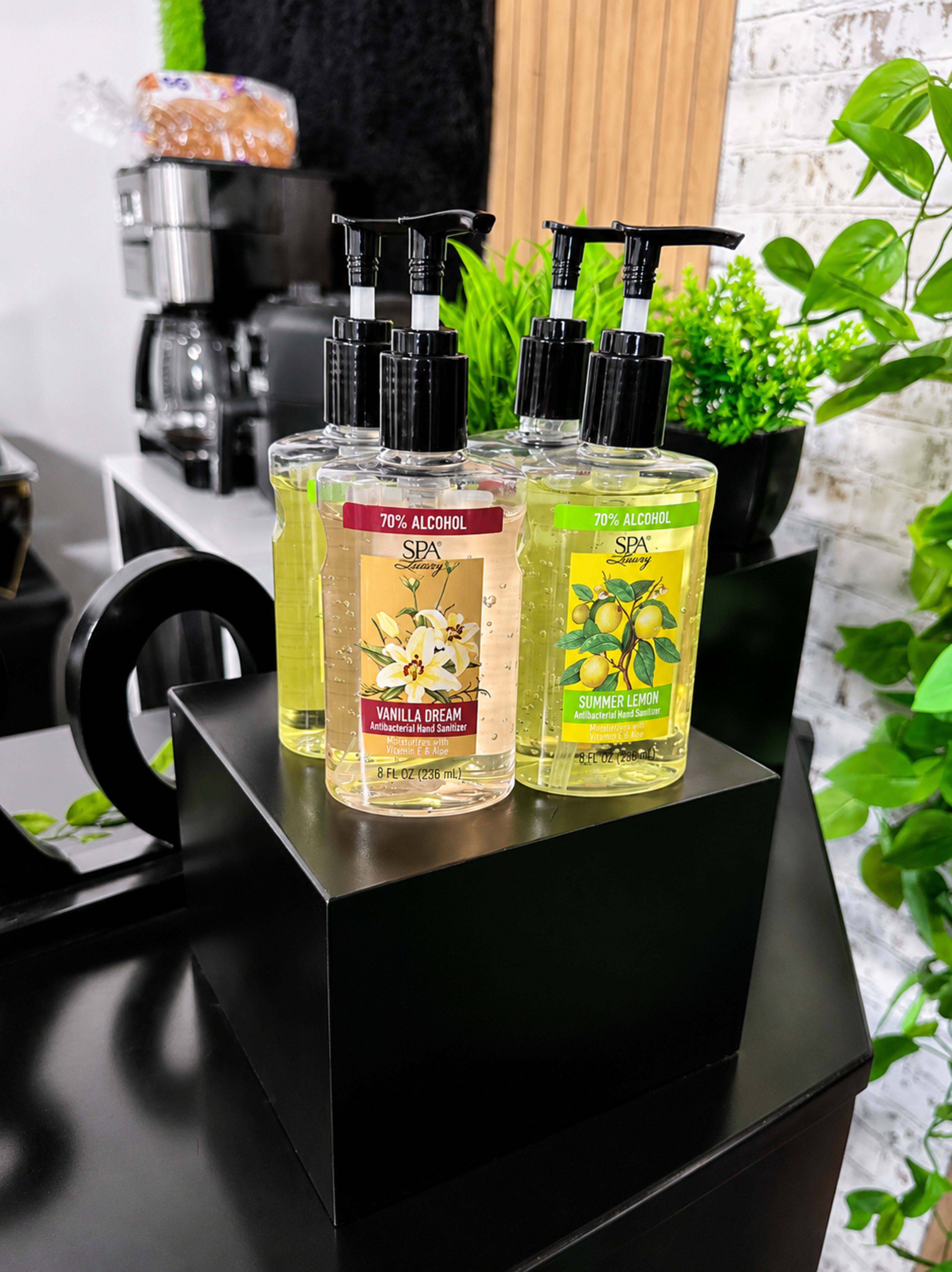 The image displays four bottles of hand sanitizers, labeled with different scents, arranged neatly on a black display stand surrounded by green plants.