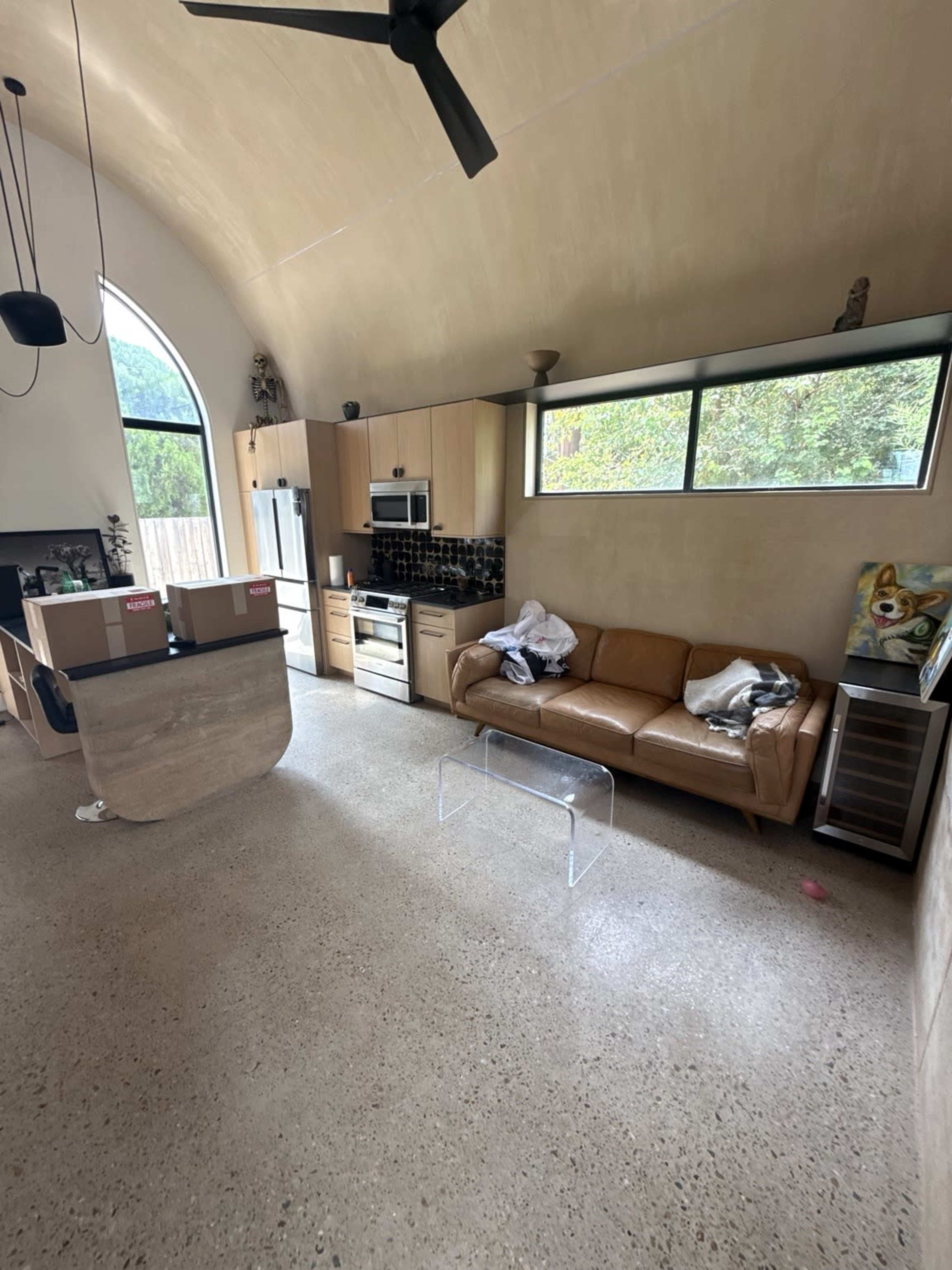 The image shows a modern kitchen and living area featuring a leather sofa, a transparent acrylic coffee table, and boxes stacked near the kitchen area.