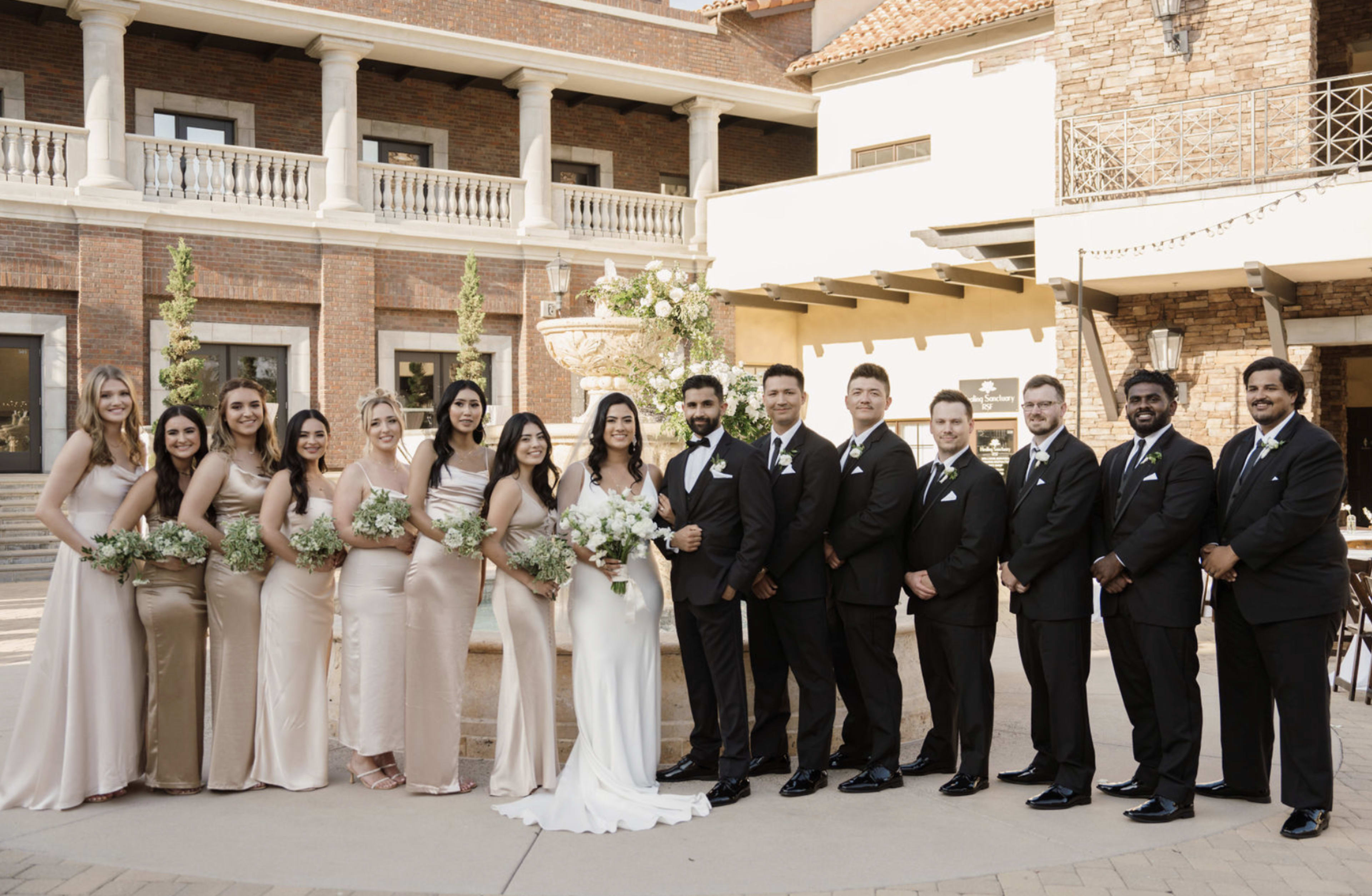 A wedding party stands together in front of a brick building with a fountain, showcasing a variety of formal attire.