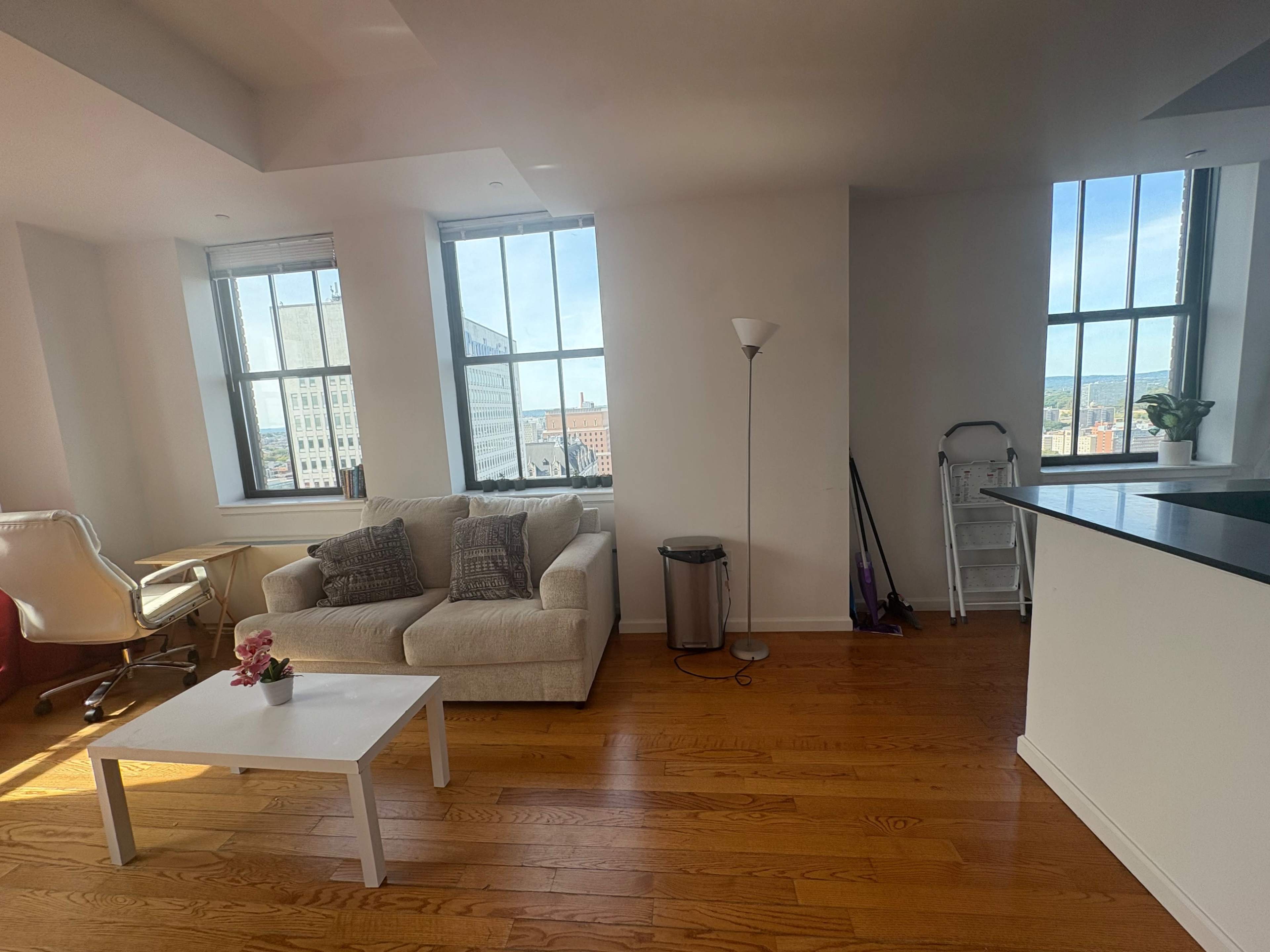 The image shows a bright, modern living room with a sofa, a small white coffee table, a standing lamp, and large windows that offer a view of the city.
