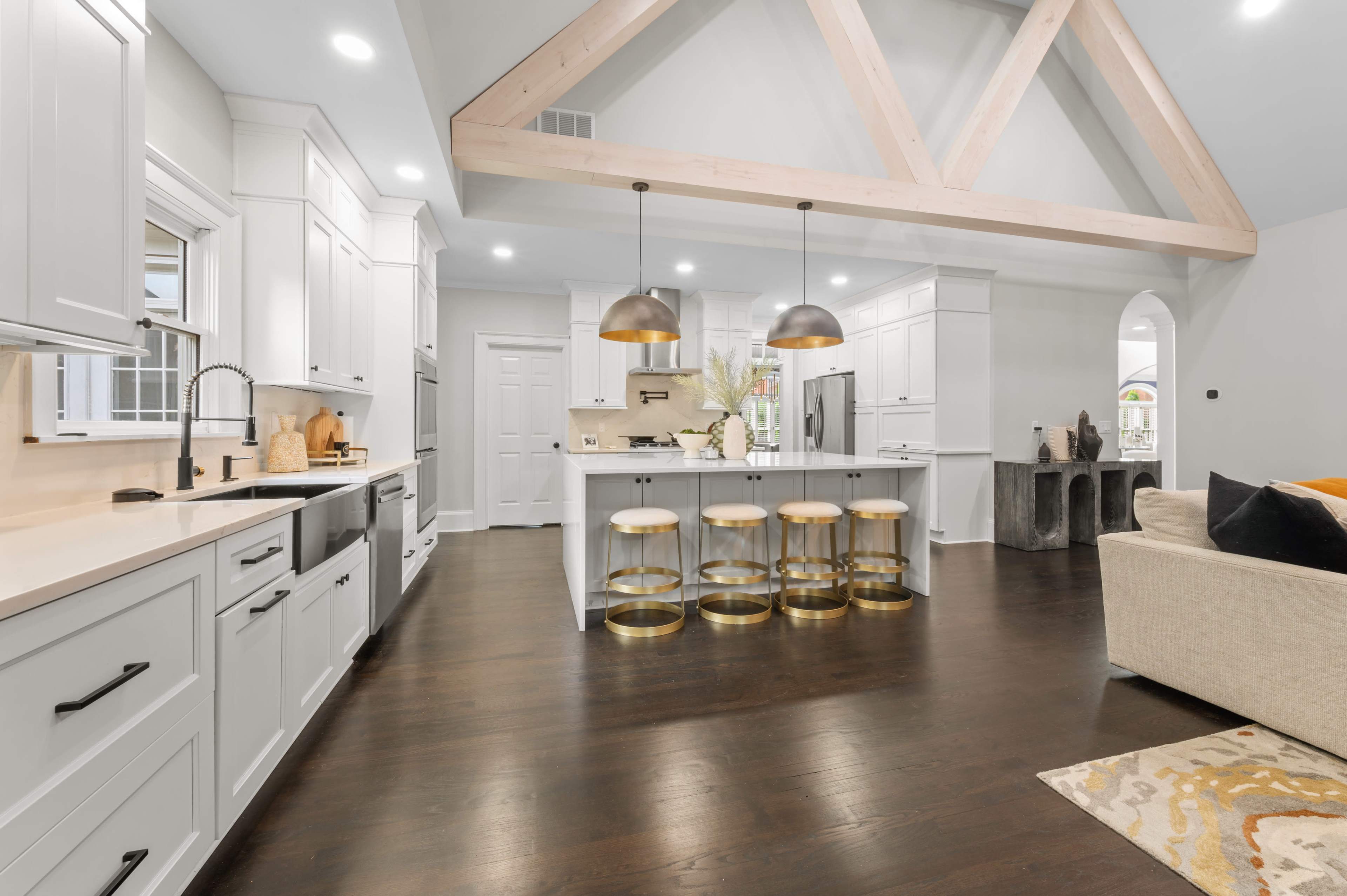 A modern kitchen with white cabinetry, a central island featuring gold-accented stools, and exposed wooden beams overhead.