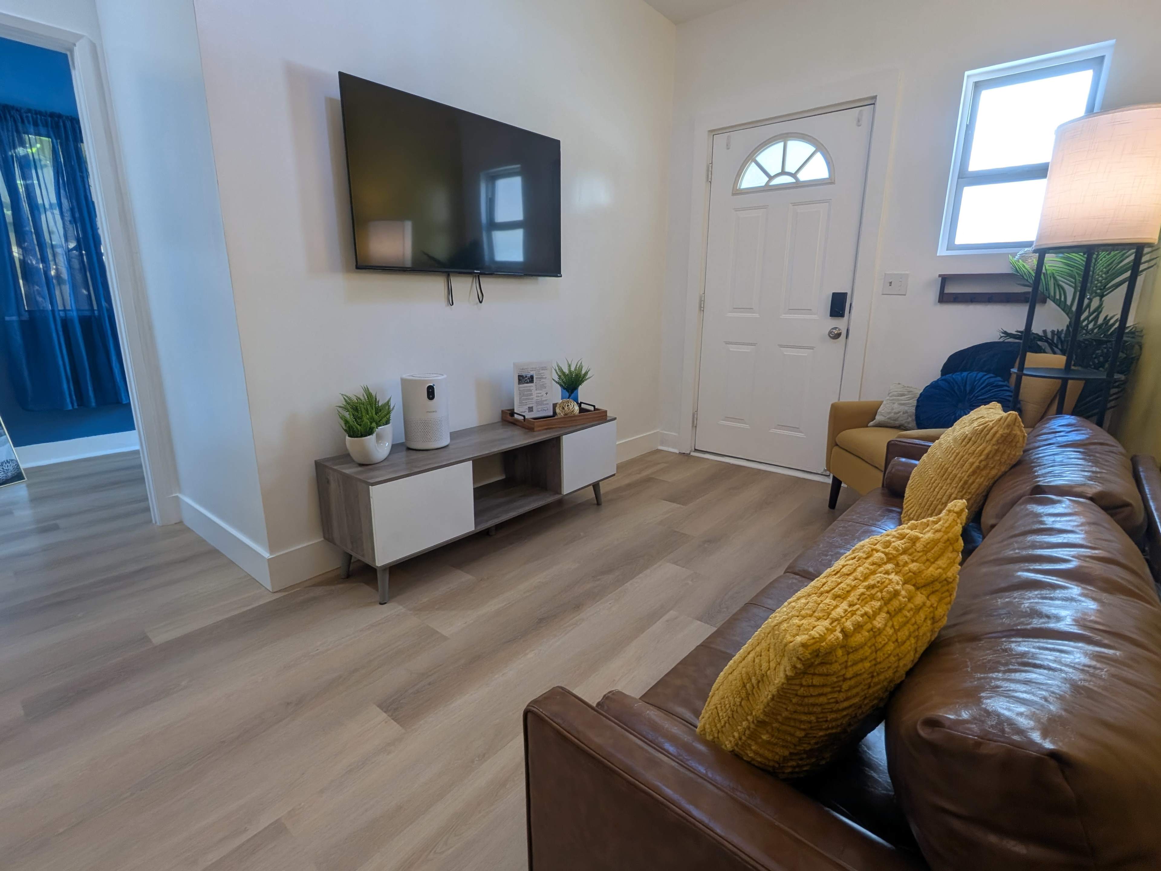 The image shows a cozy living room with a large television mounted on the wall, a brown leather sofa with yellow pillows, and a white door leading to the outside.