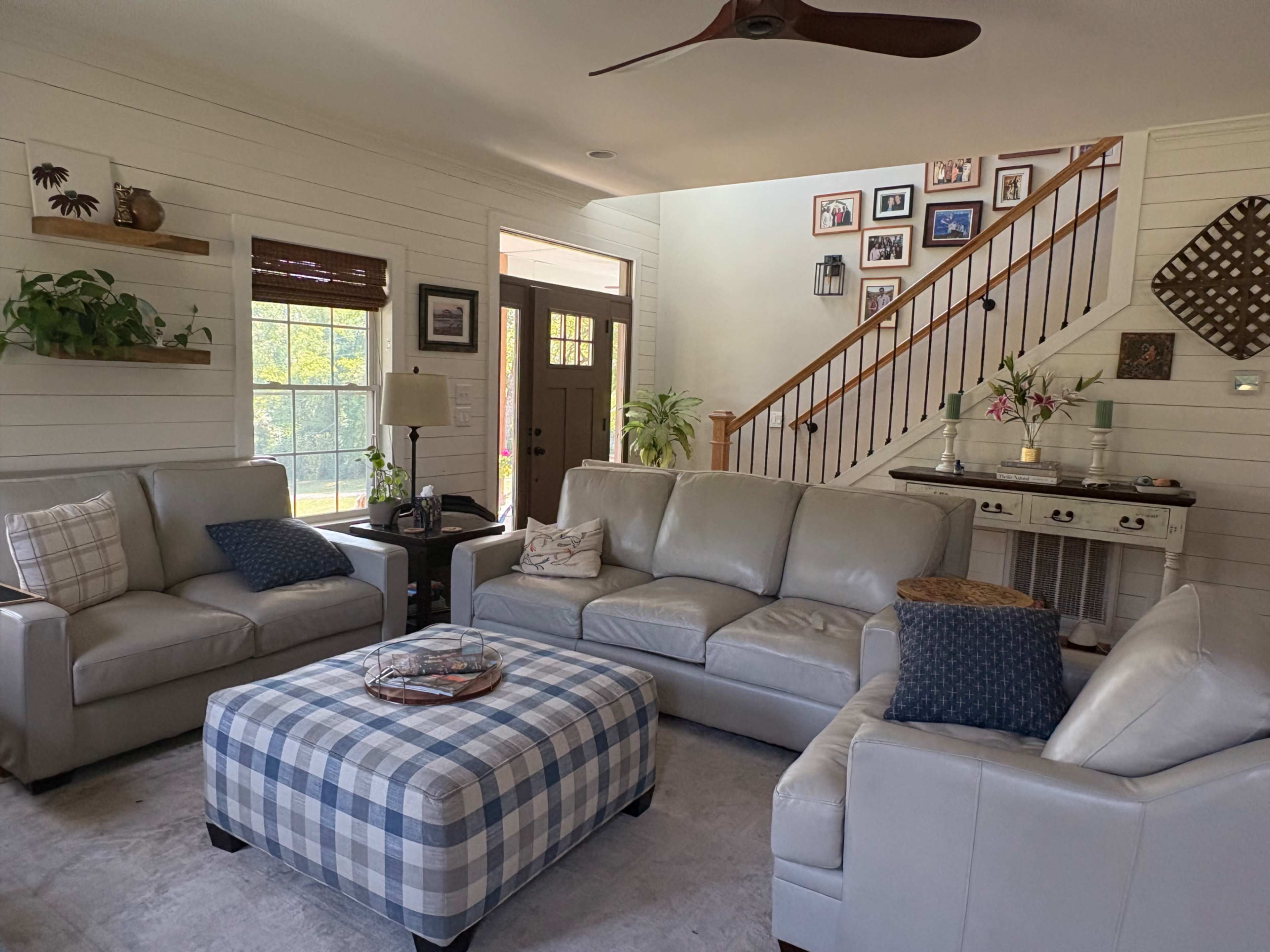 The image shows a bright, modern living room with a light-colored leather sofa, a checkered ottoman, and a staircase adorned with framed photos.