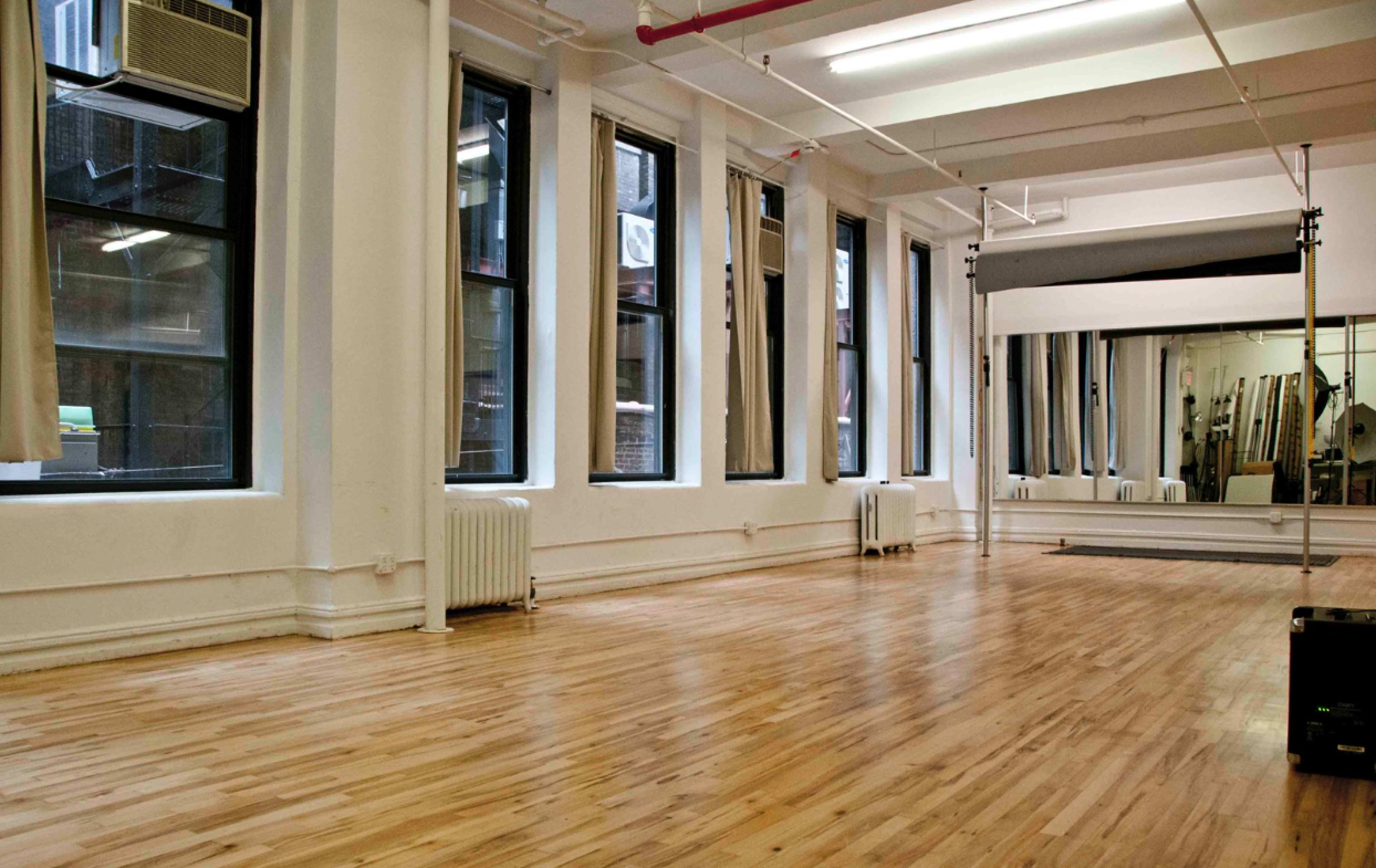 The image shows a spacious, empty studio with large windows, wooden flooring, and minimal furnishings.