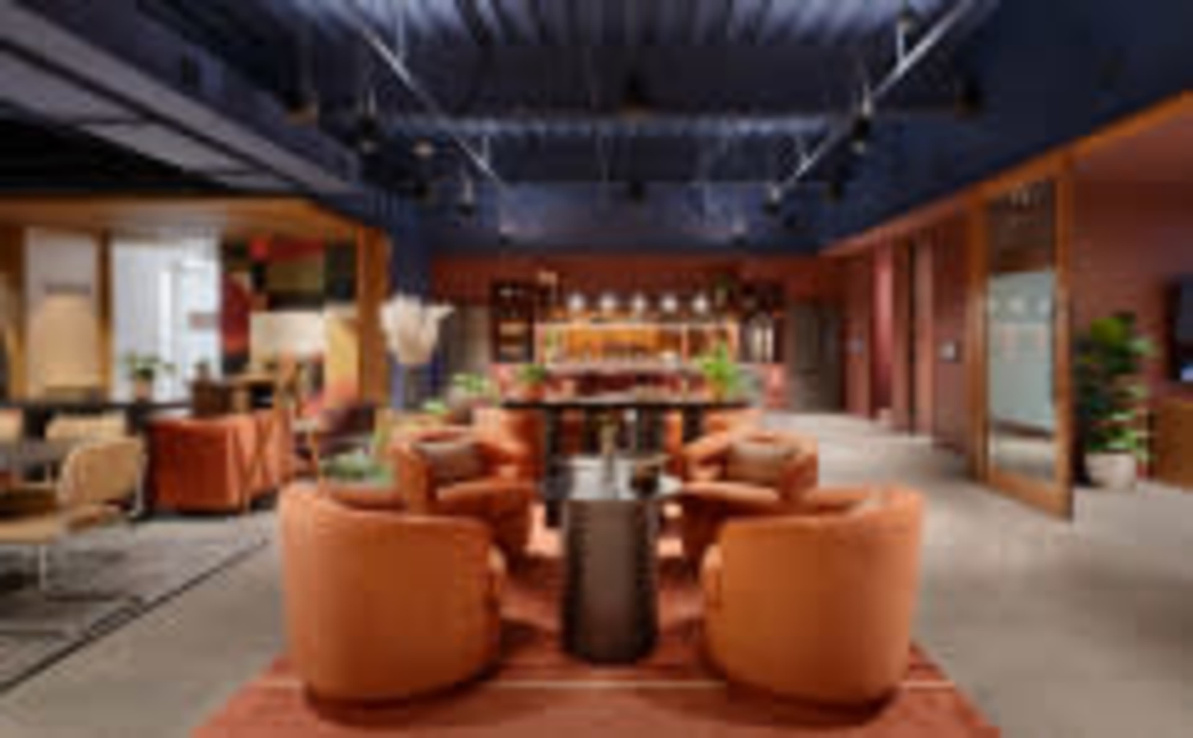 The image shows a modern lounge area featuring orange chairs arranged around a circular table, surrounded by plants and shelves in a spacious room.