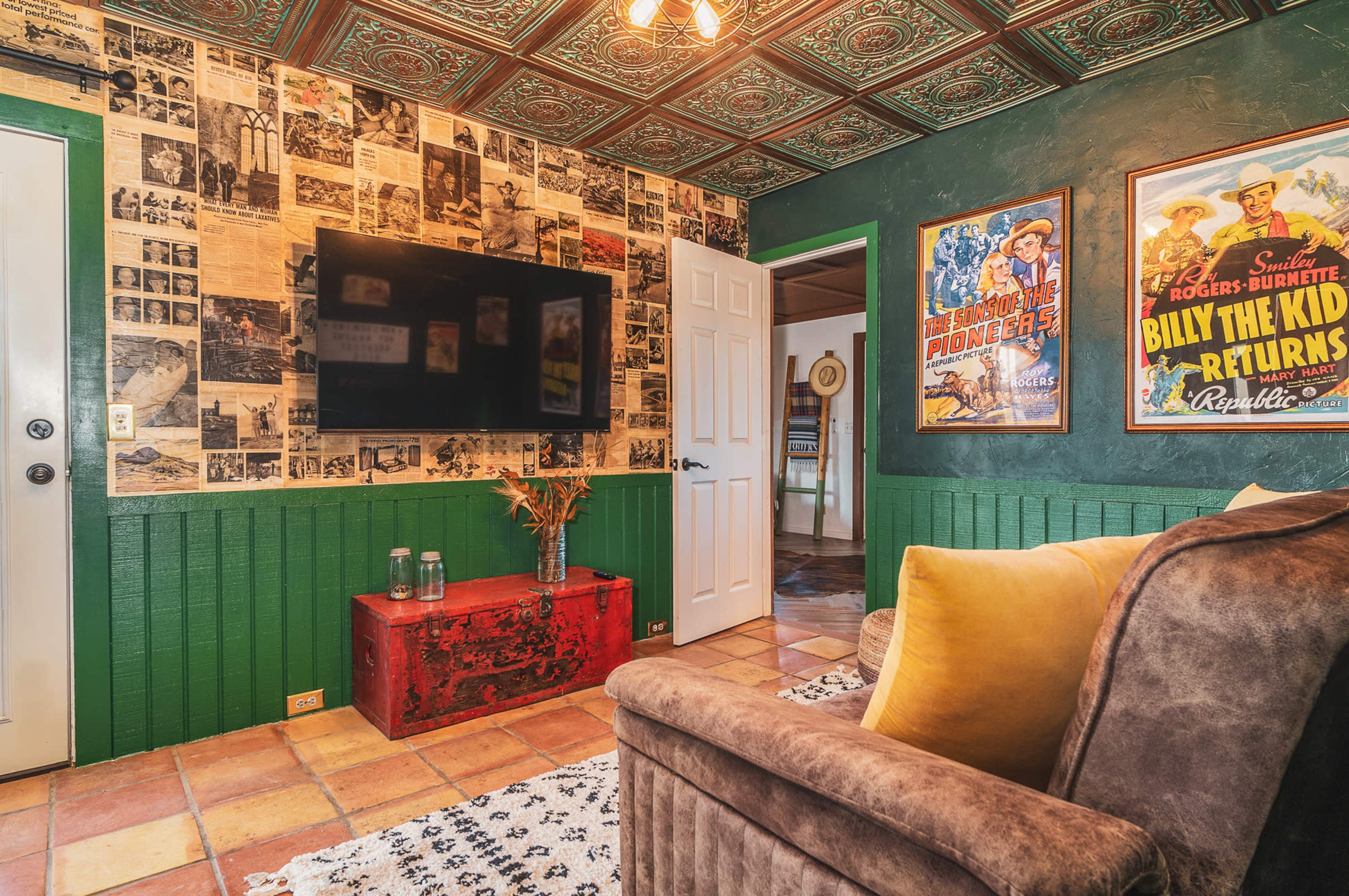 The room features a wall adorned with vintage newspaper clippings and posters, a television mounted on the wall, and a cozy seating area with a patterned rug.