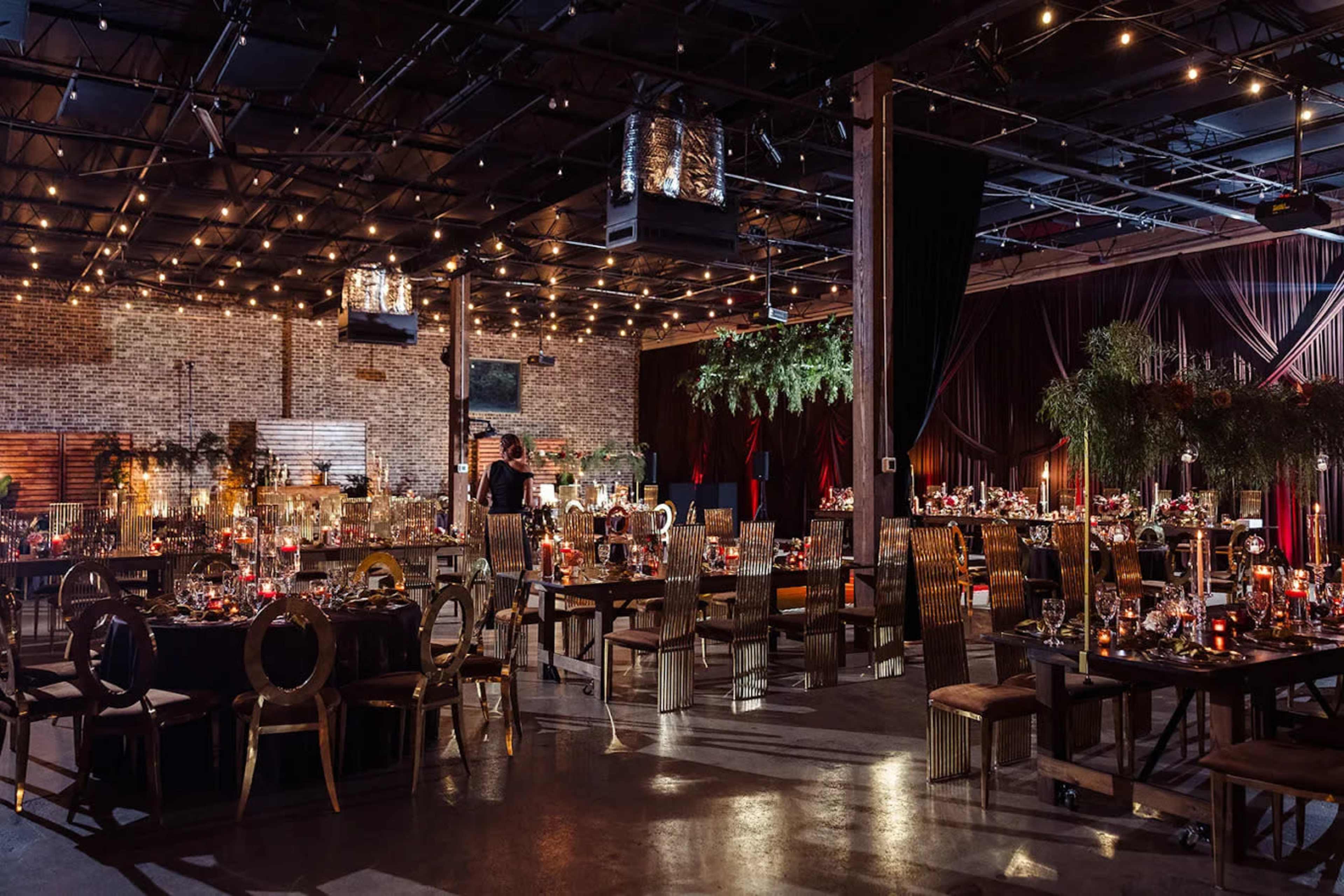 The image shows a spacious event venue filled with elegantly arranged tables, ambient lighting, and decorative elements, creating a festive atmosphere.