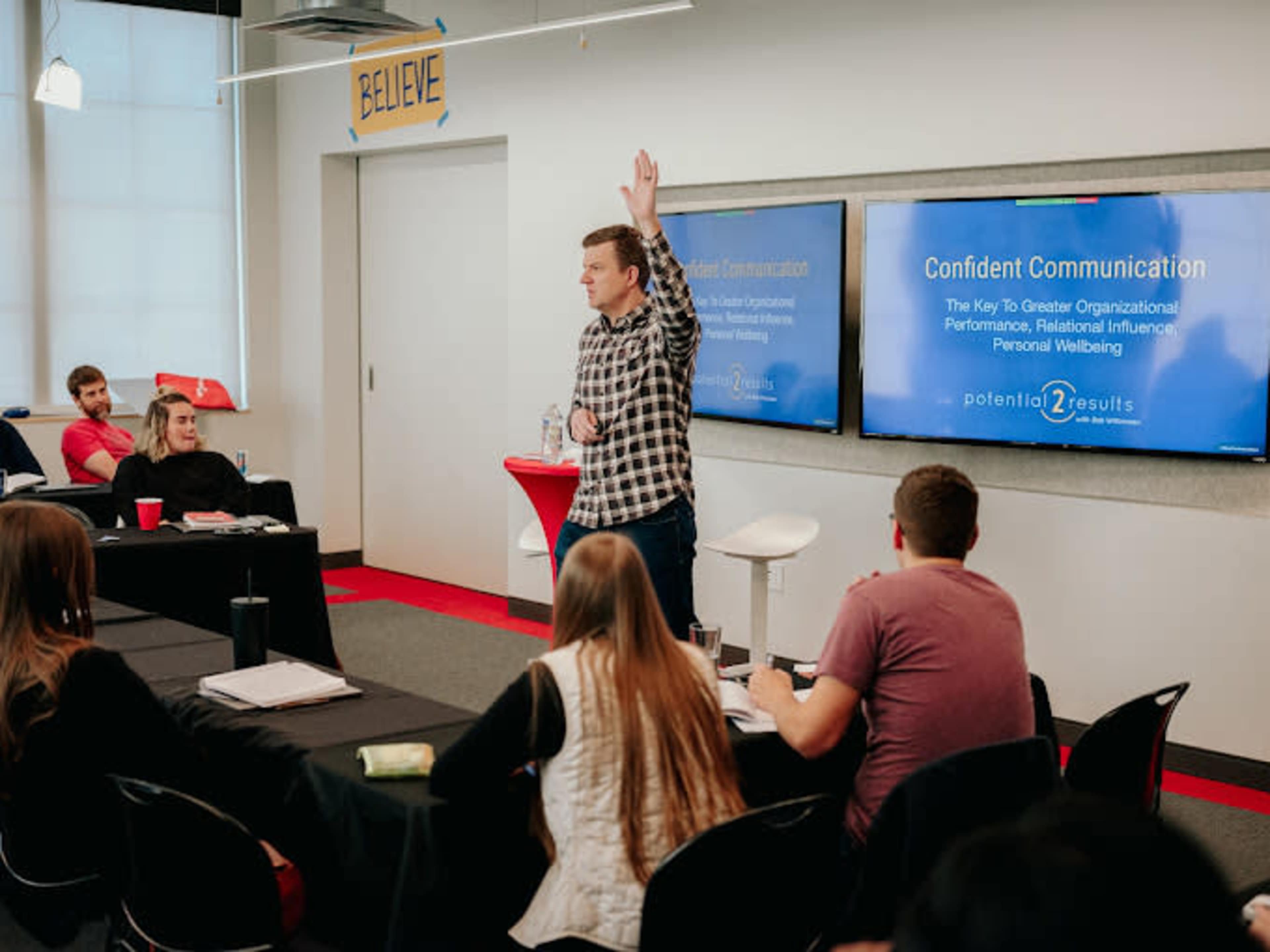 A presenter stands at the front of a classroom, gesturing while discussing "Confident Communication" to an audience seated at tables.