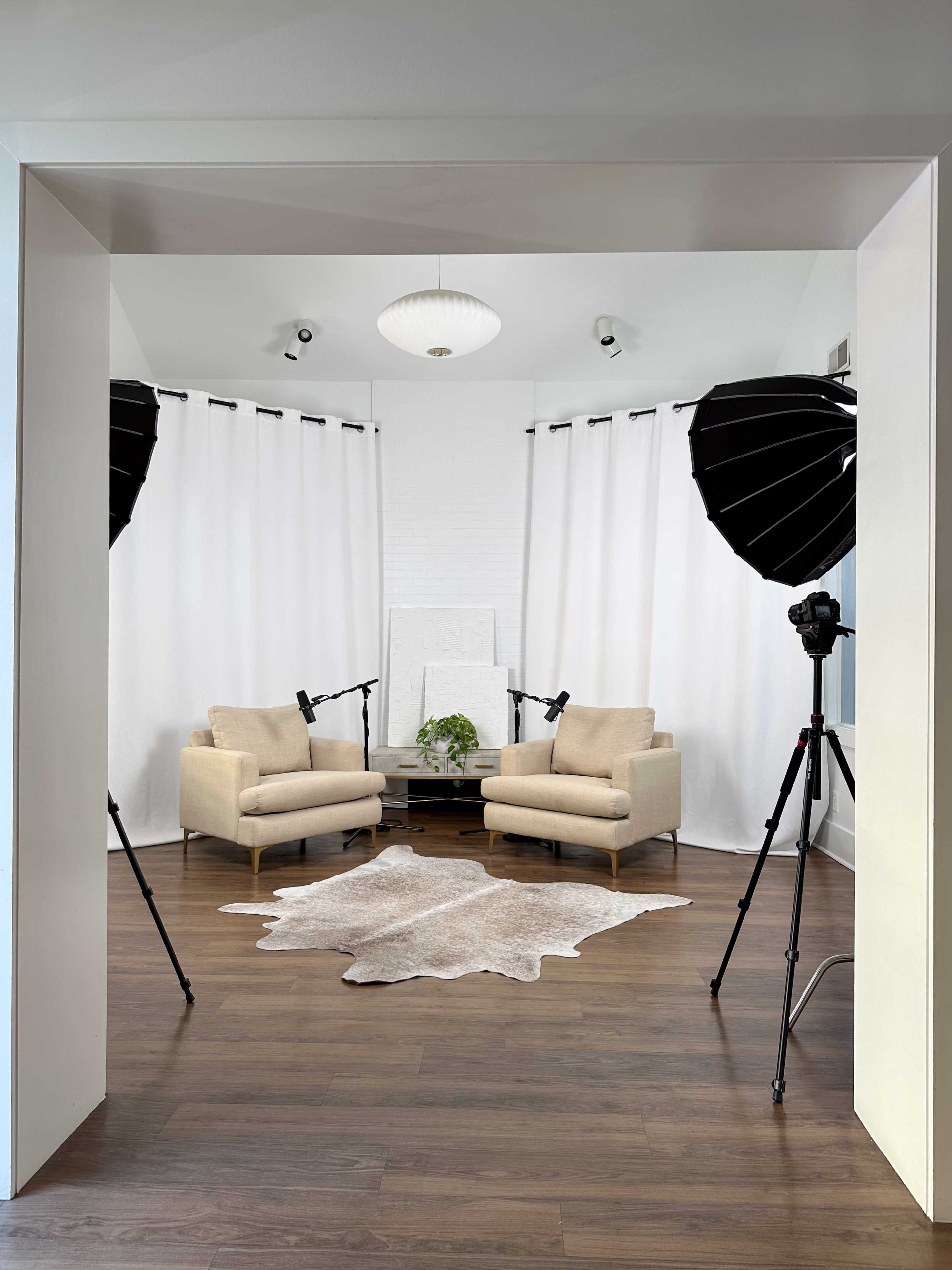 The image features a neatly arranged studio setup with two chairs flanked by softbox lights, a rug on the floor, and white curtains creating a backdrop.