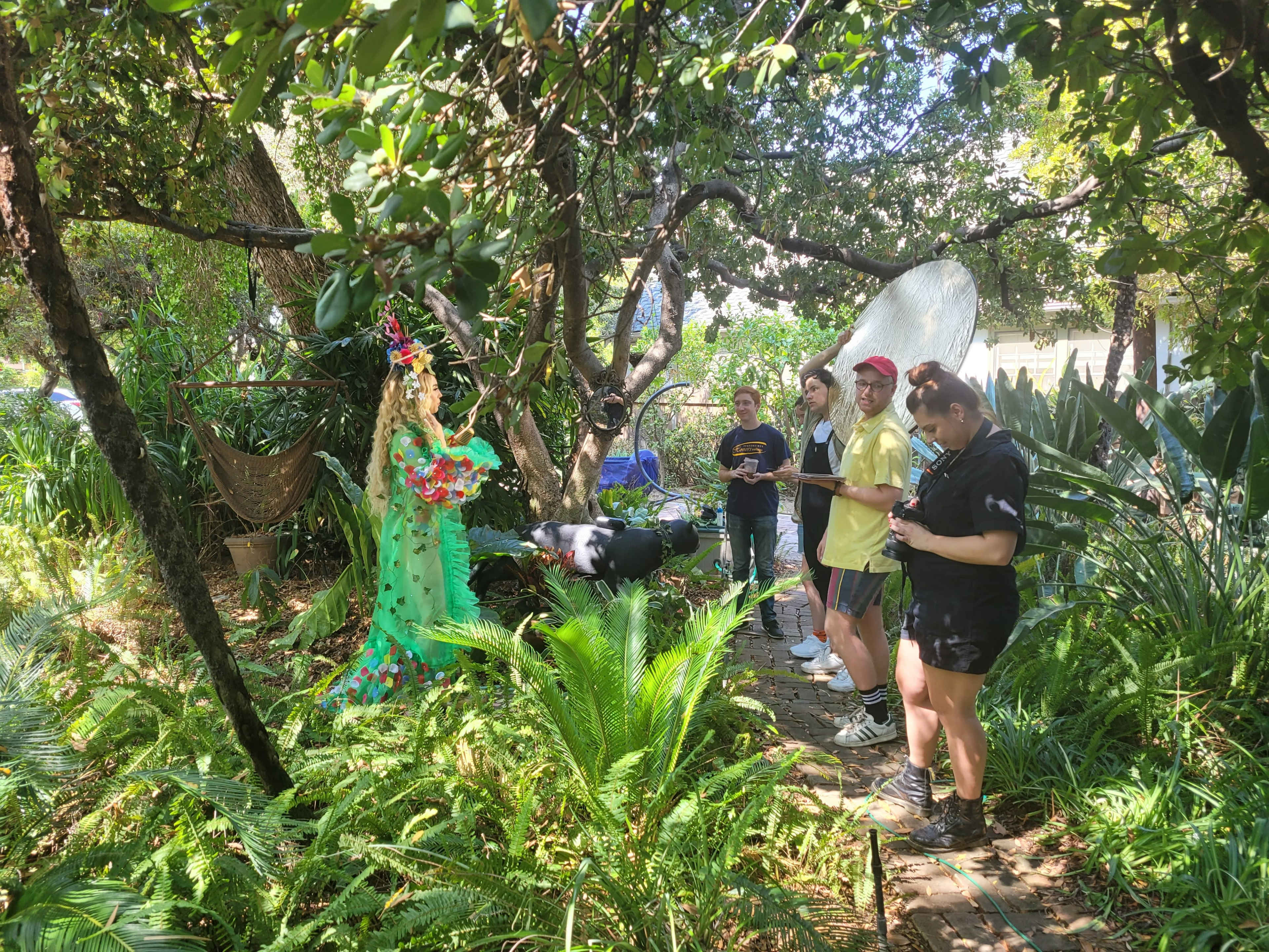 A group of people stands on a wooden path in a lush garden while observing a figure dressed in a colorful costume among the greenery.