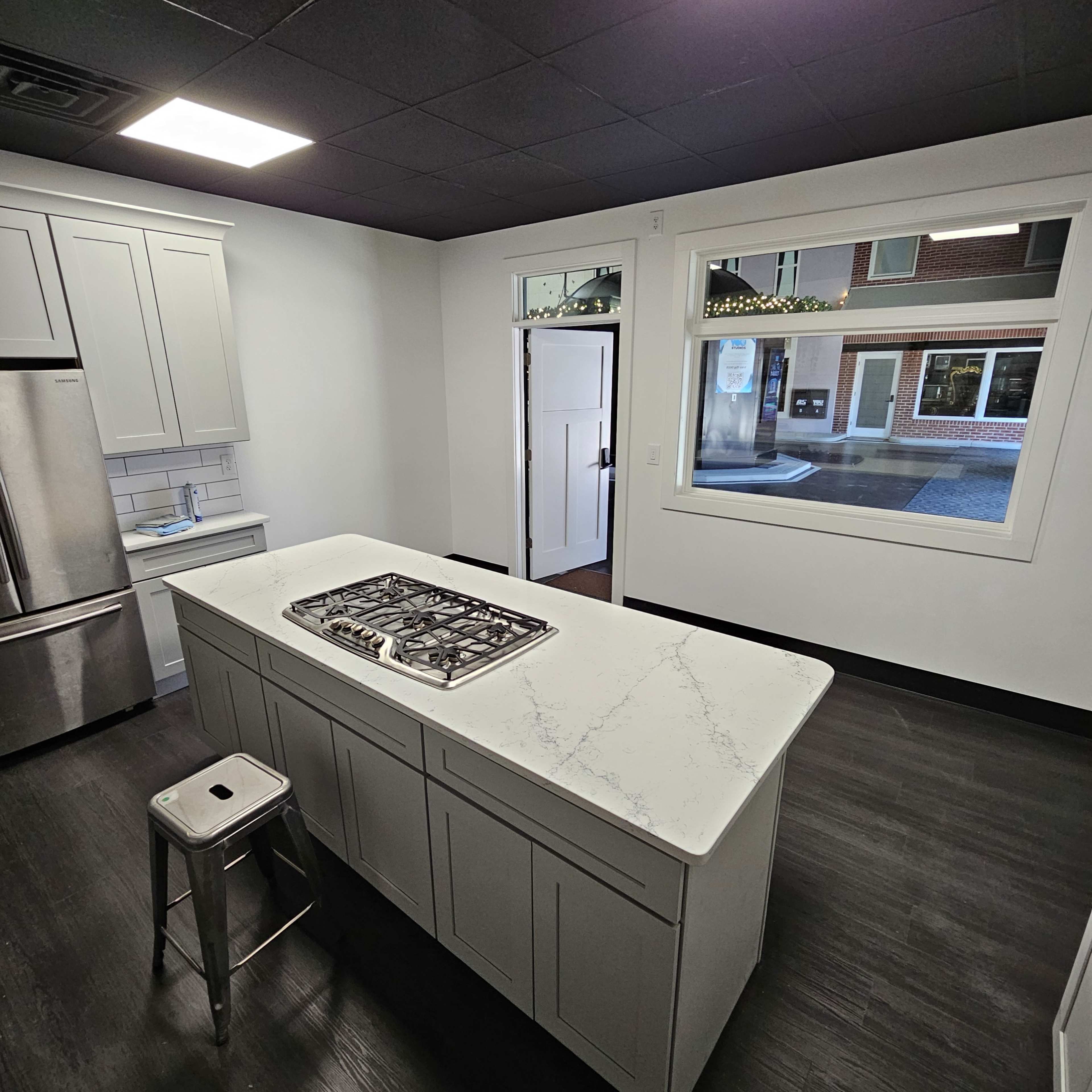 The image shows a modern kitchen featuring a central island with a gas stovetop, light gray cabinetry, and a large window overlooking a street.