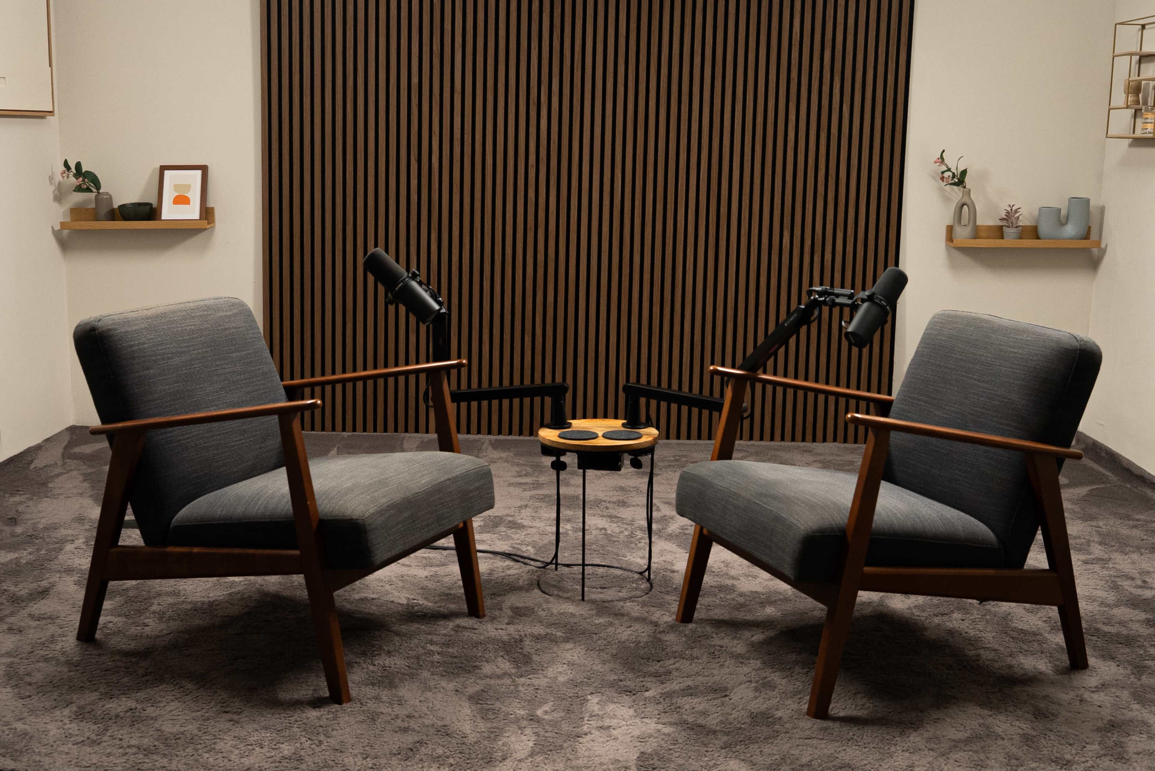 The image shows two mid-century modern armchairs facing each other, with a small round table between them, set in a room with a textured wall and decorative shelves.