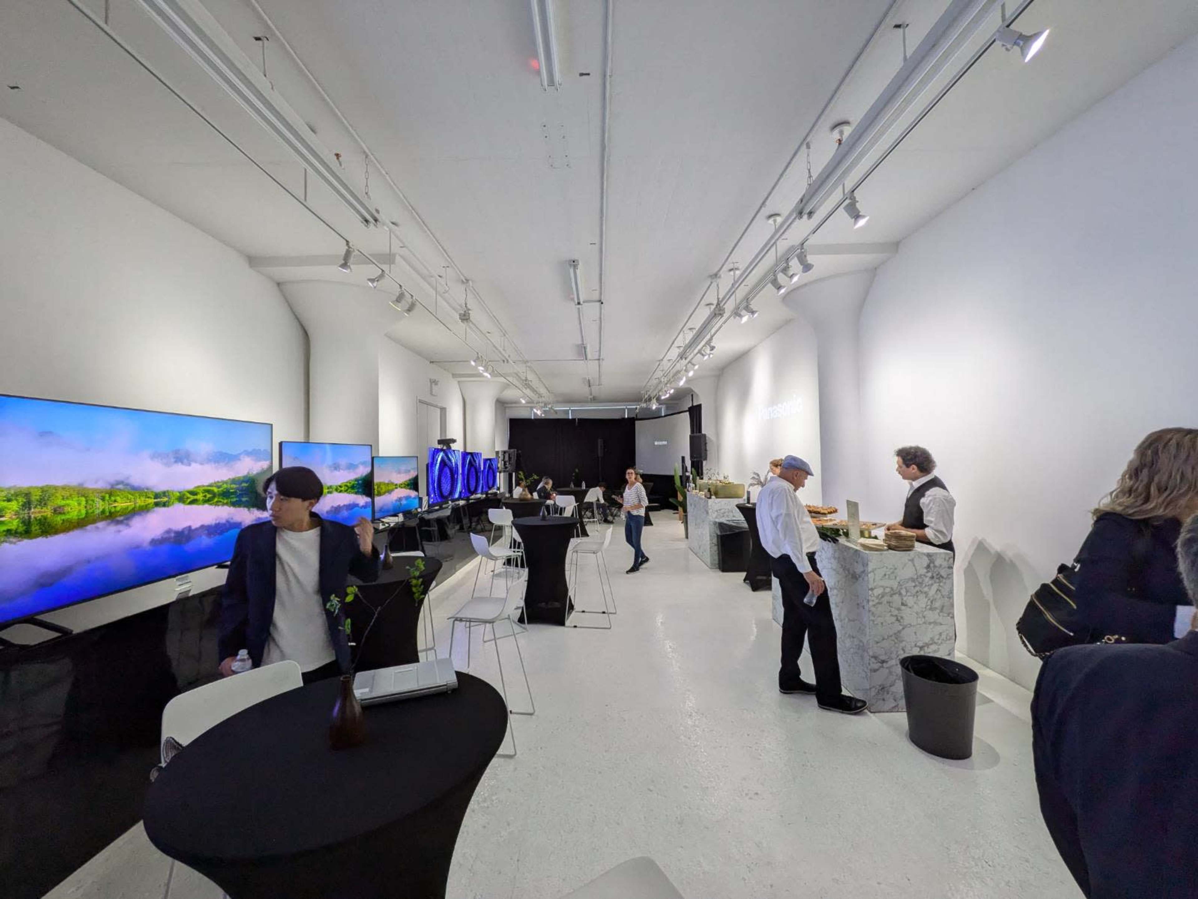 The scene shows an exhibition space featuring large screens displaying nature images, with people interacting and a catering setup along one side.