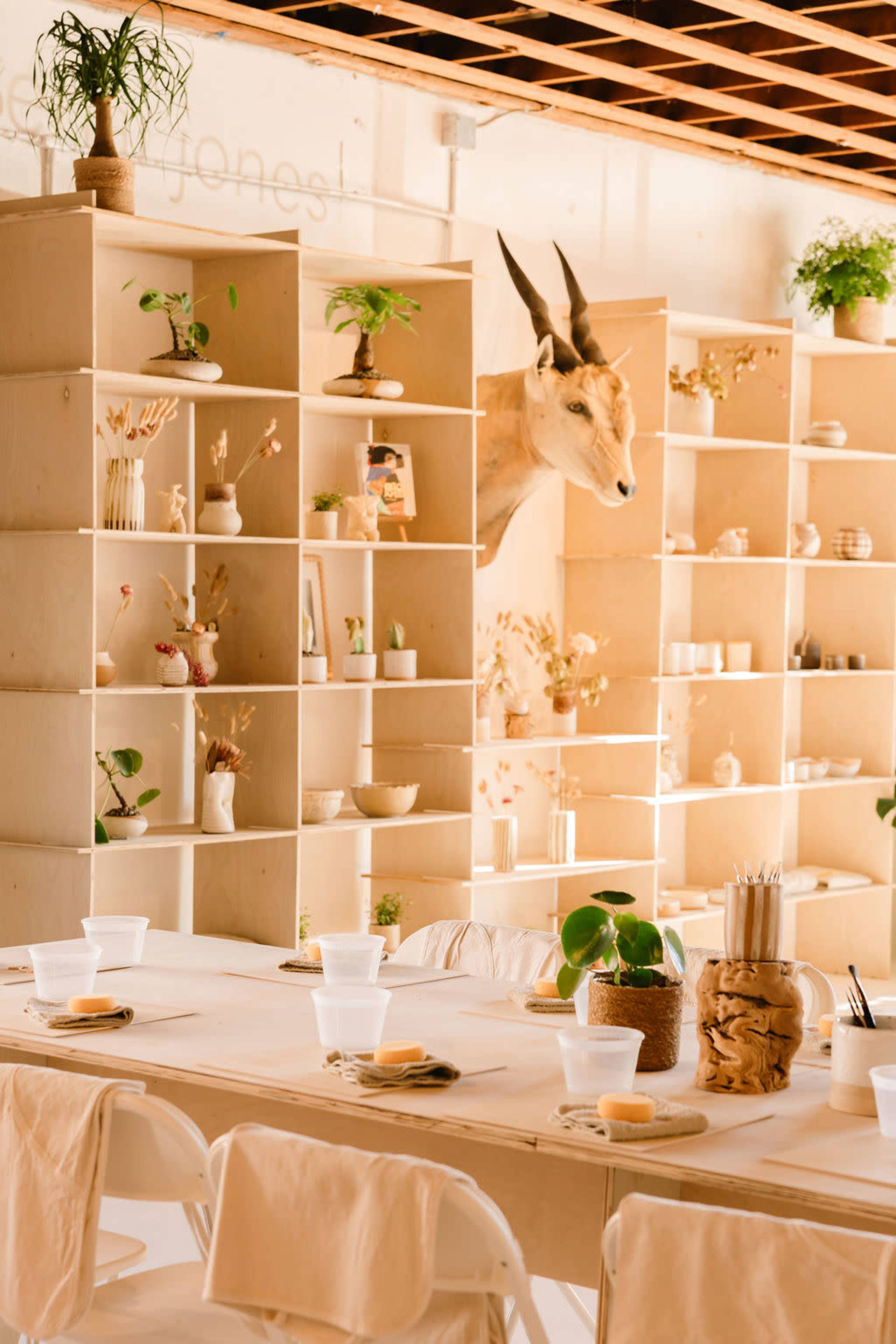 The image shows a minimalistic interior of a room with light-colored wooden shelves filled with decorative items, and a table set for a gathering with plastic cups and table settings.