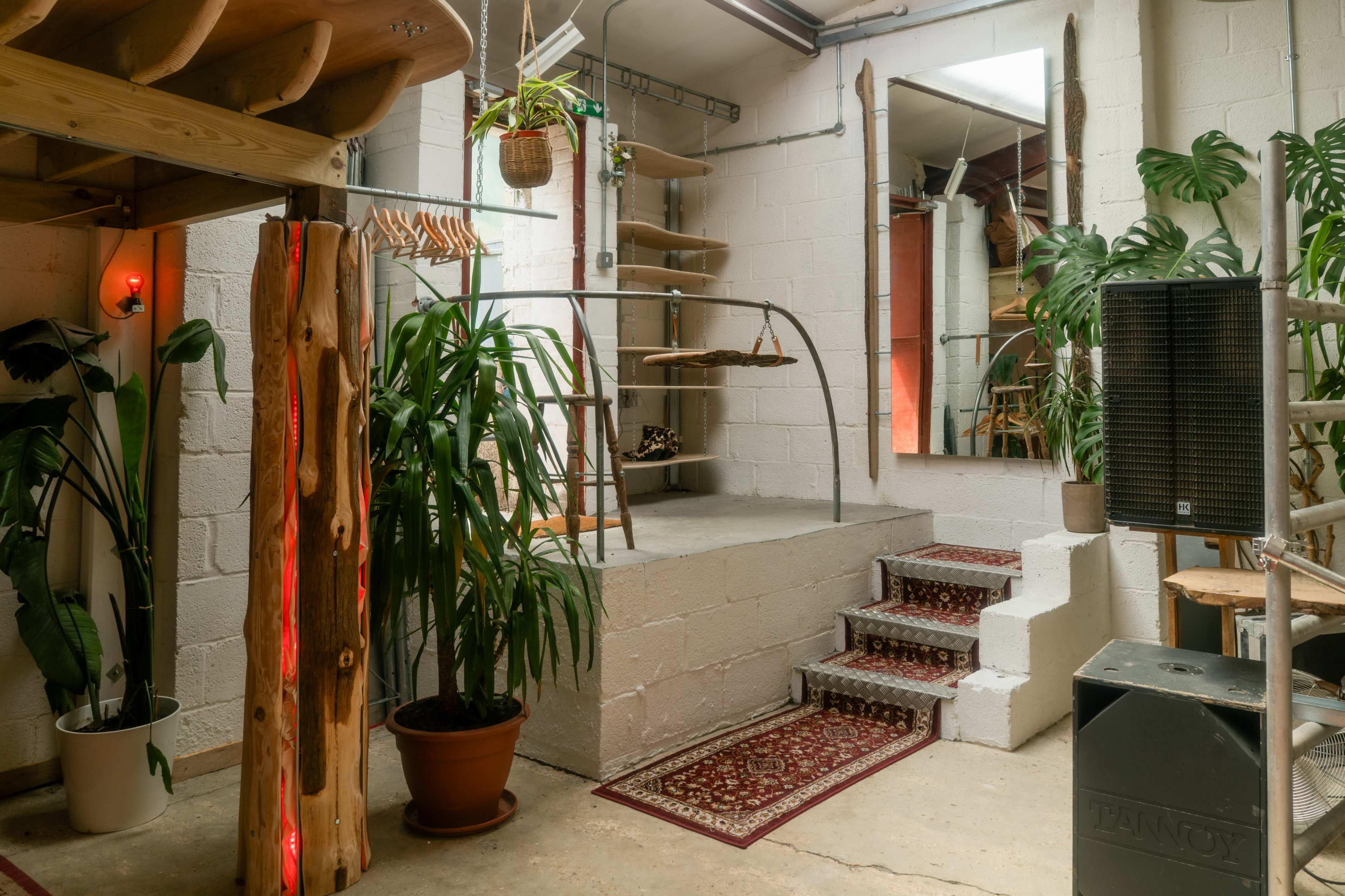 The image shows an interior space featuring a staircase, shelves, potted plants, and a mirror, with a mix of natural wood elements and concrete walls.