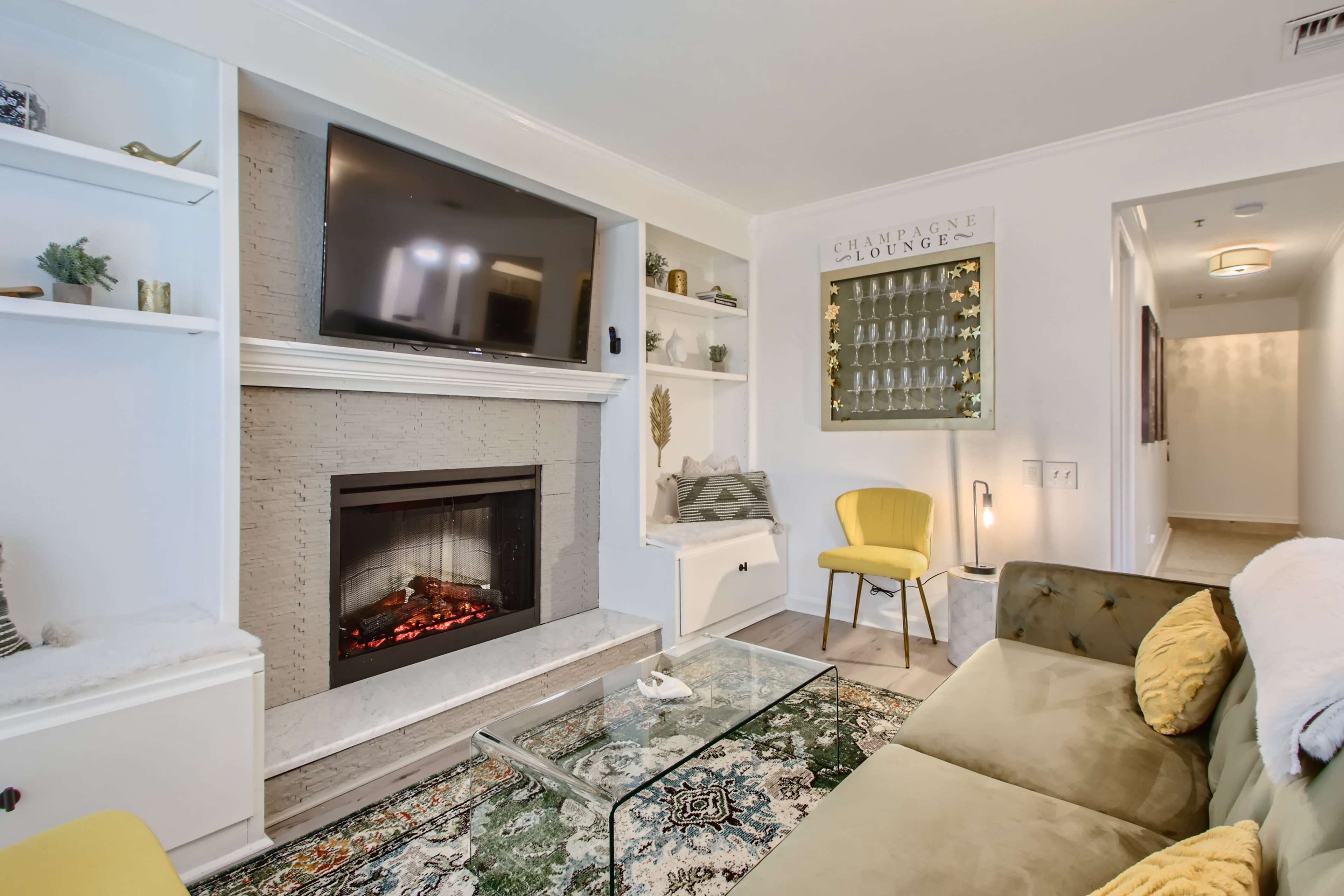 The living room features a modern fireplace beneath a wall-mounted TV, with a yellow chair, a glass coffee table, and decorative shelves.