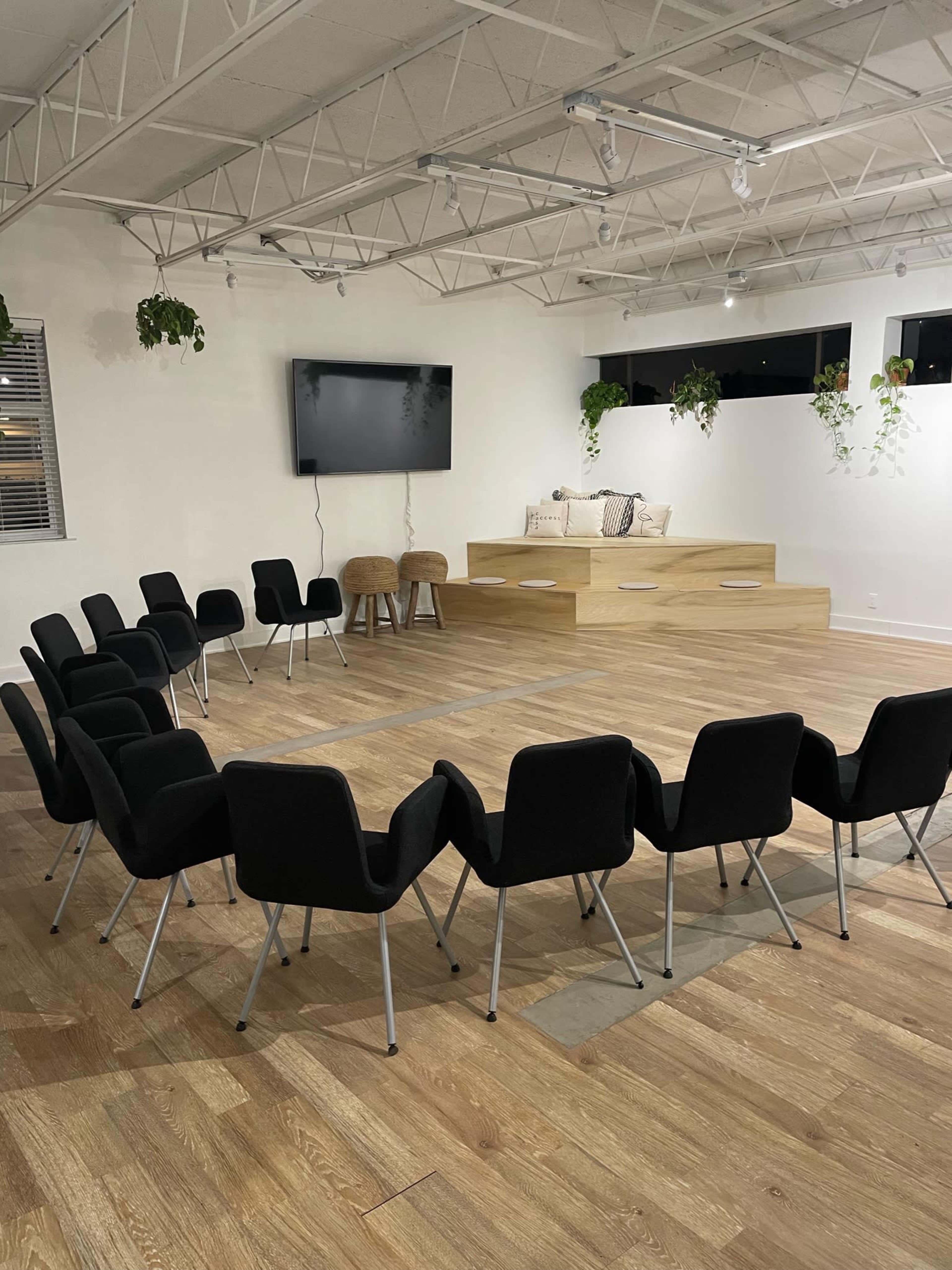 A circular arrangement of black chairs is set up in a brightly lit room with a large screen and a seating area against the wall.