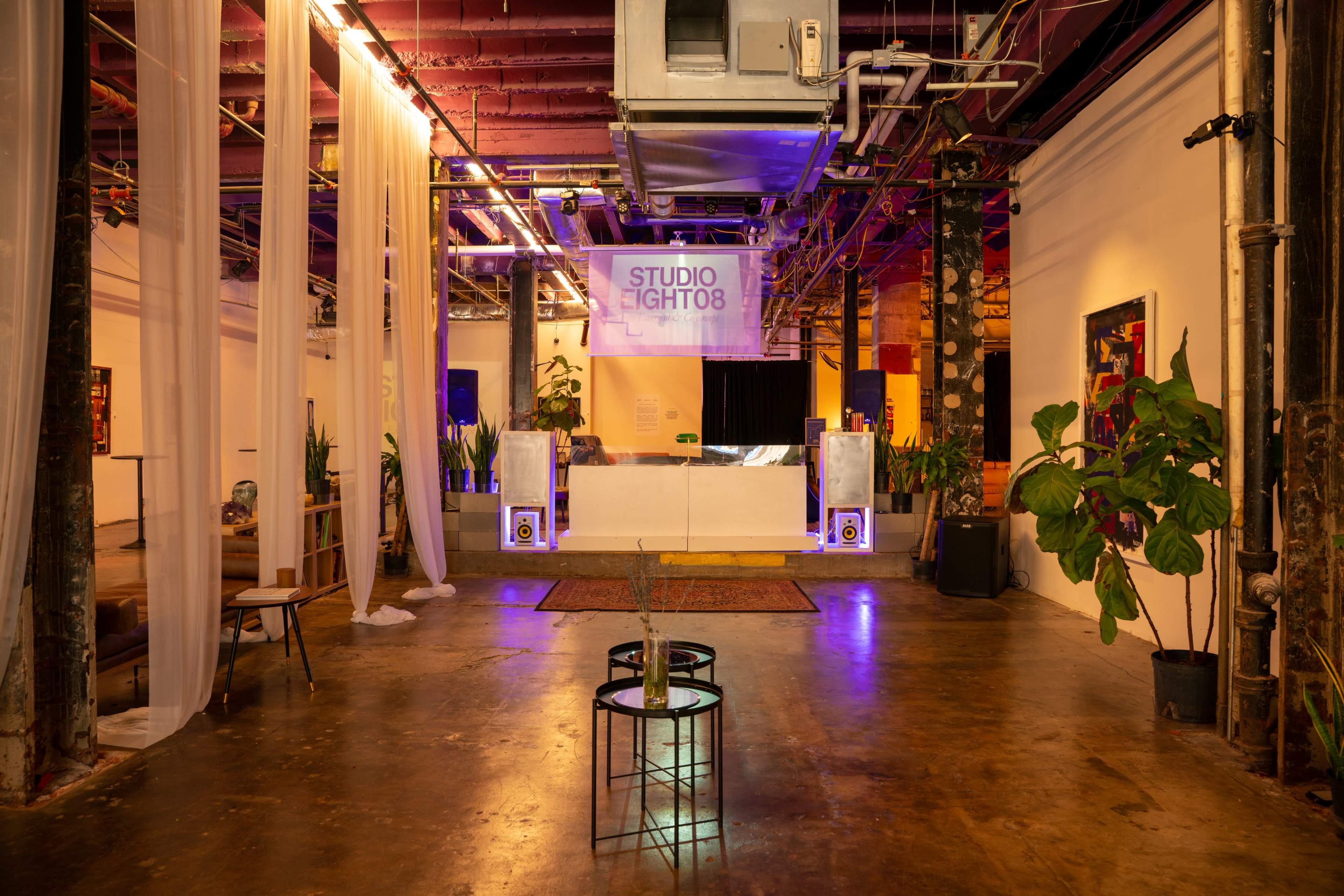 The image depicts an industrial event space with exposed pipes and colorful lighting, featuring a seating area, a stage, and decorative plants.