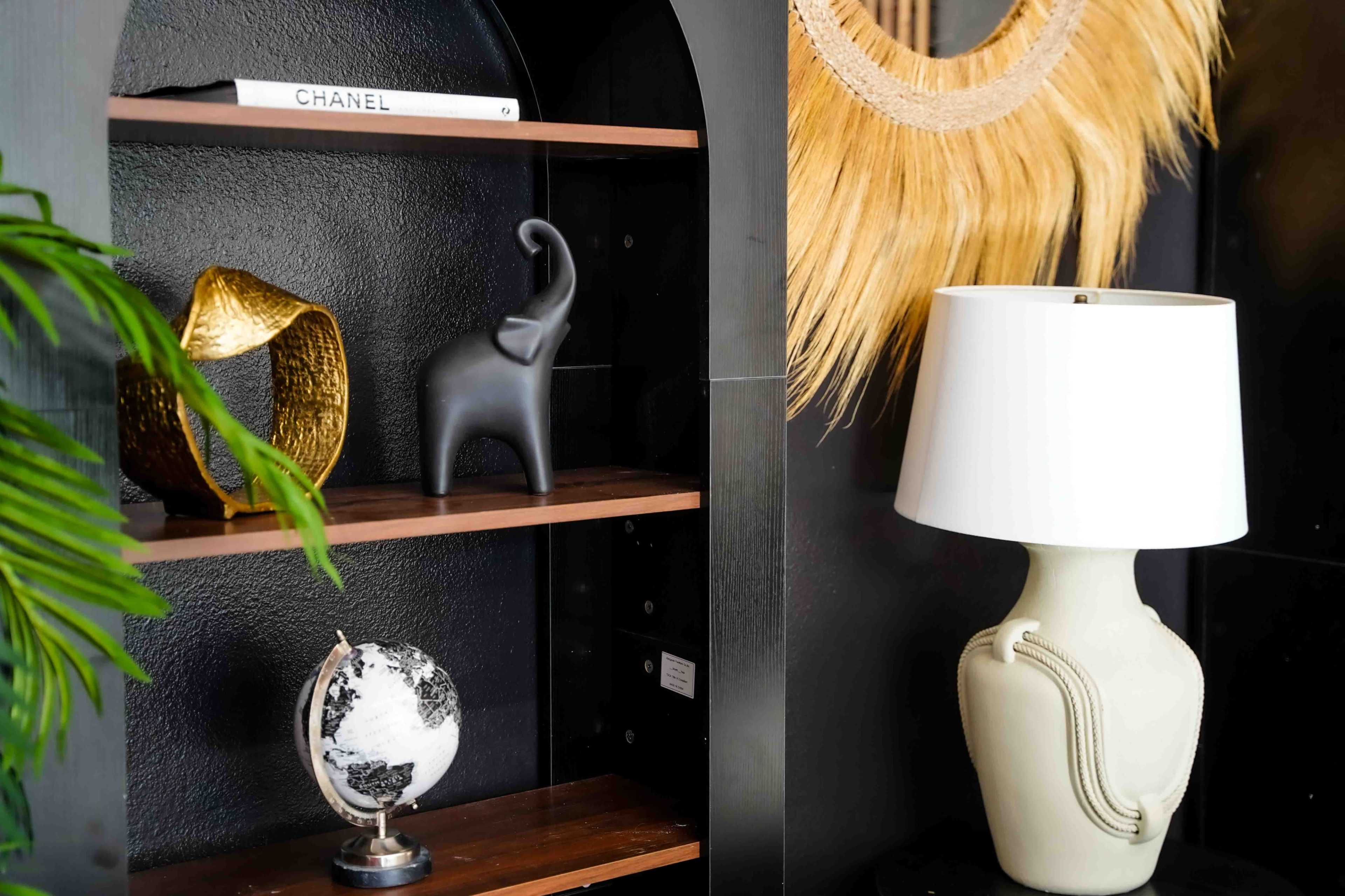 The image features a dark shelf displaying a decorative elephant figurine, a gold sculpture, a globe, and a lamp, with a large wall-mounted decorative piece made of natural fibers in the background.