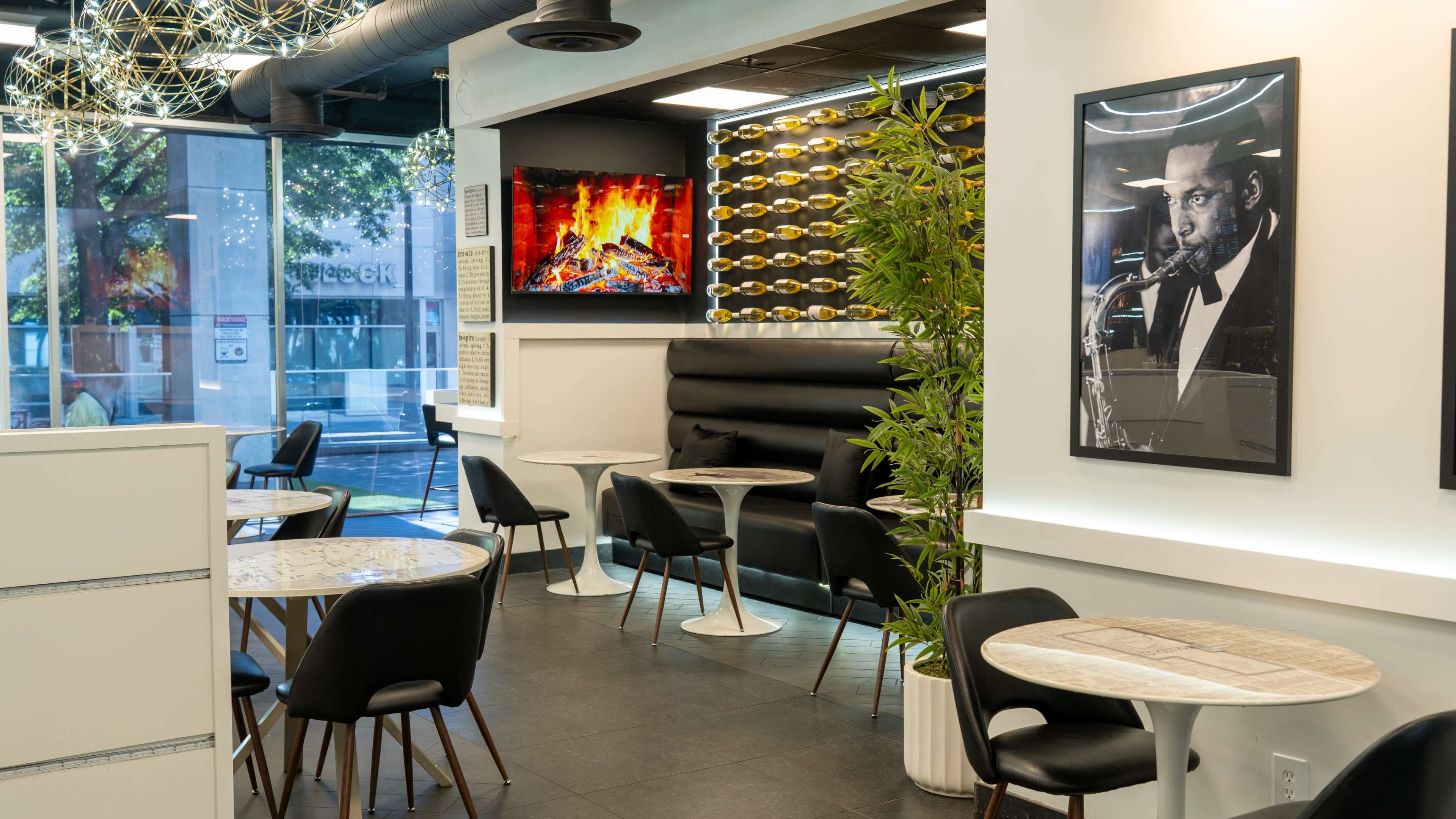 The image shows a modern café interior featuring sleek tables and chairs, a black leather bench, a wall with decorative elements, and a screen displaying a fireplace.