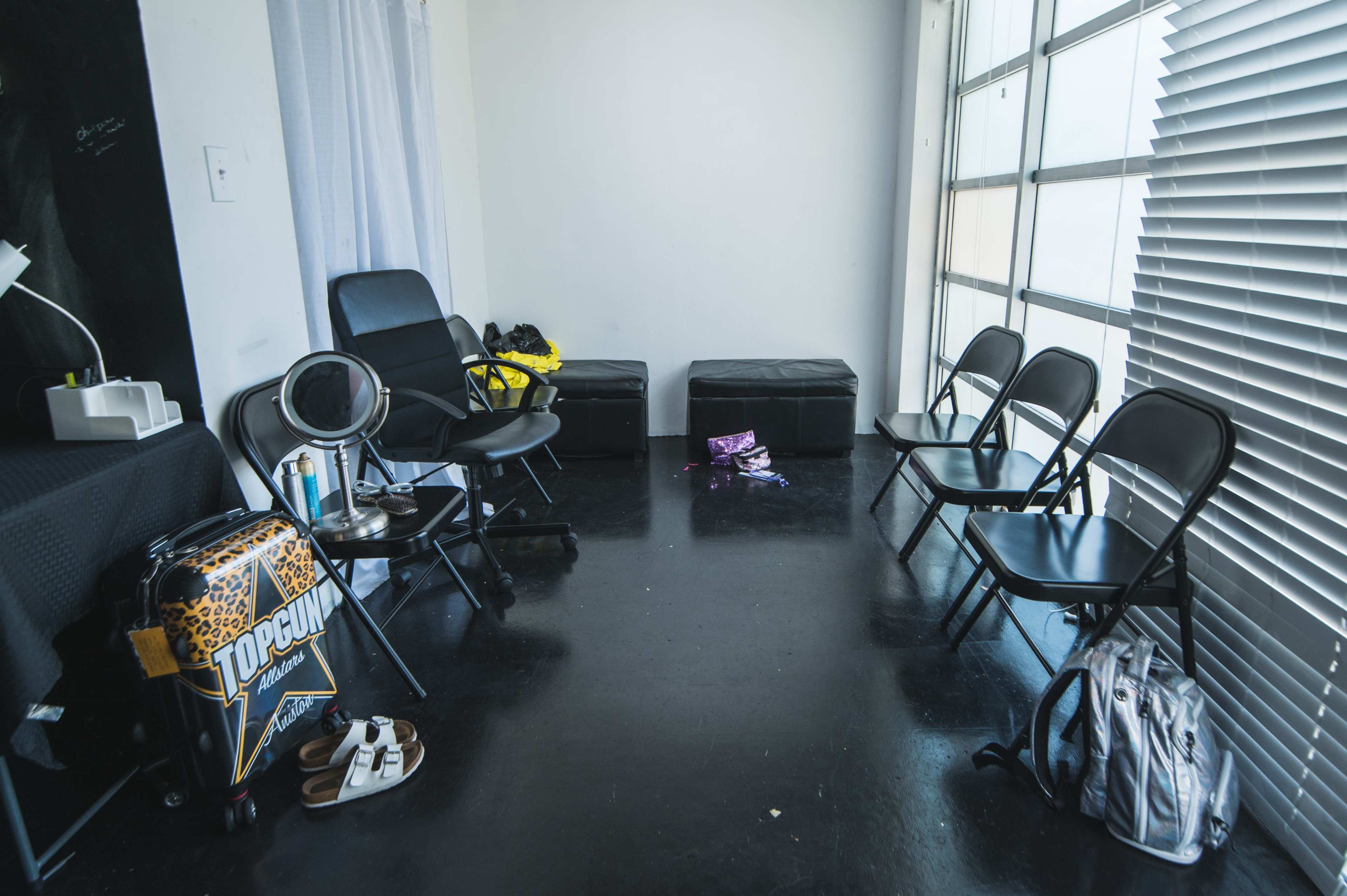 A sparsely furnished room with several folding chairs, a small table, and storage ottomans, illuminated by natural light from a large window.