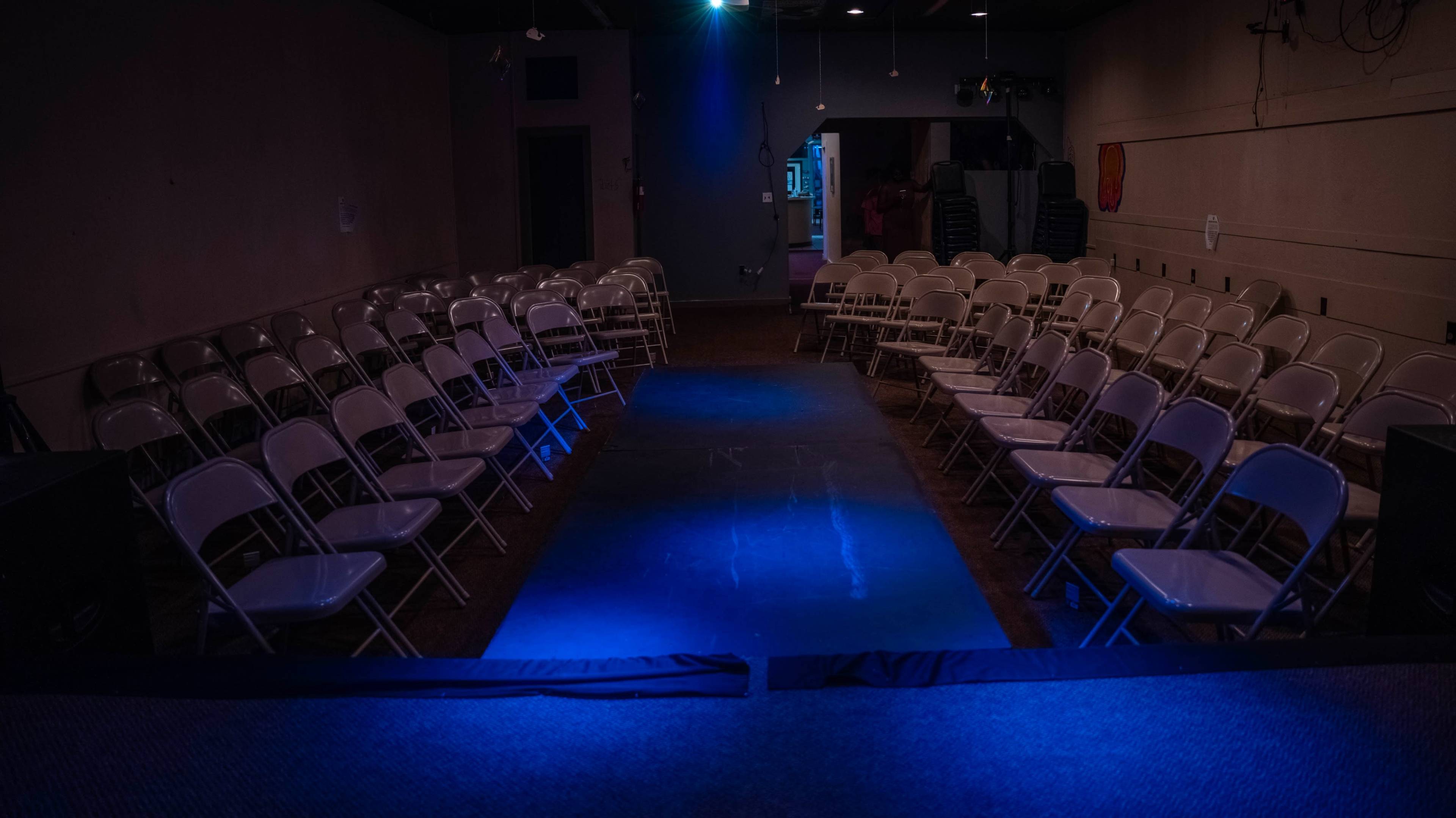A dimly lit indoor venue features rows of white folding chairs facing a central runway illuminated by a blue light.