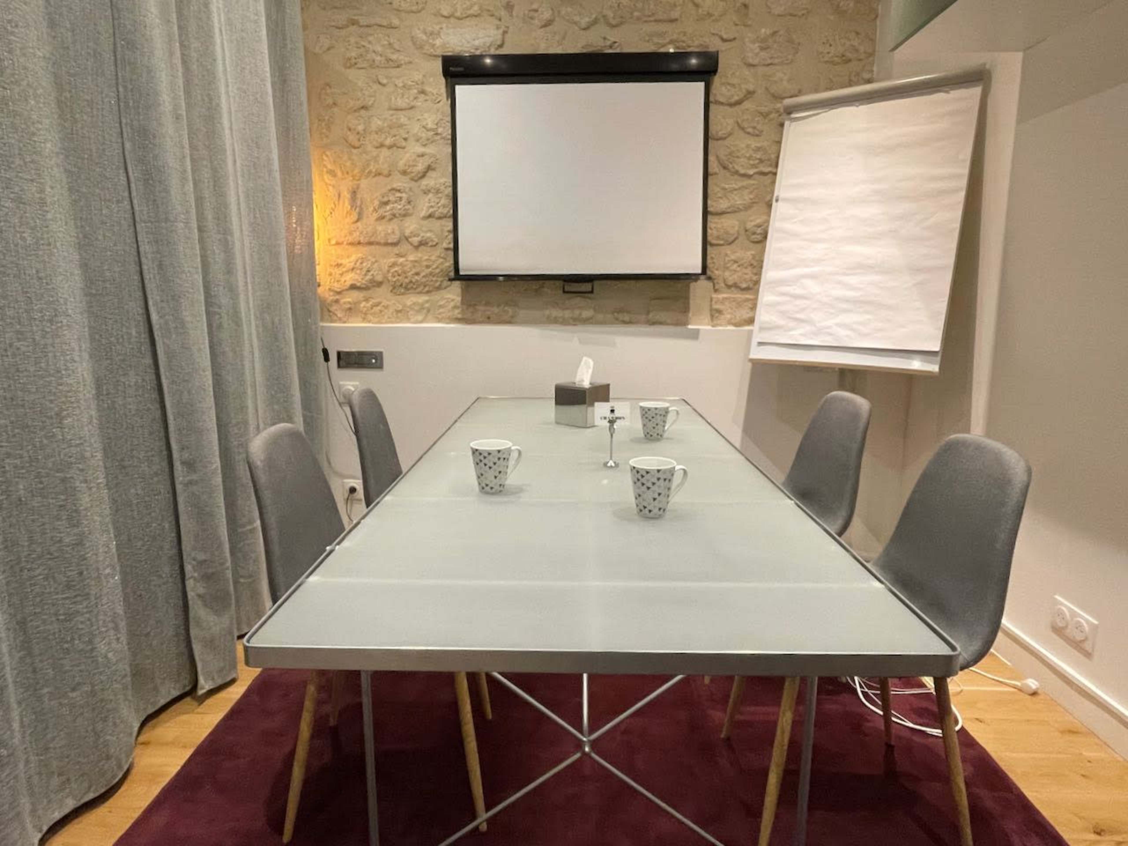 A modern meeting room with a glass table, four gray chairs, a projector screen, and a flip chart.
