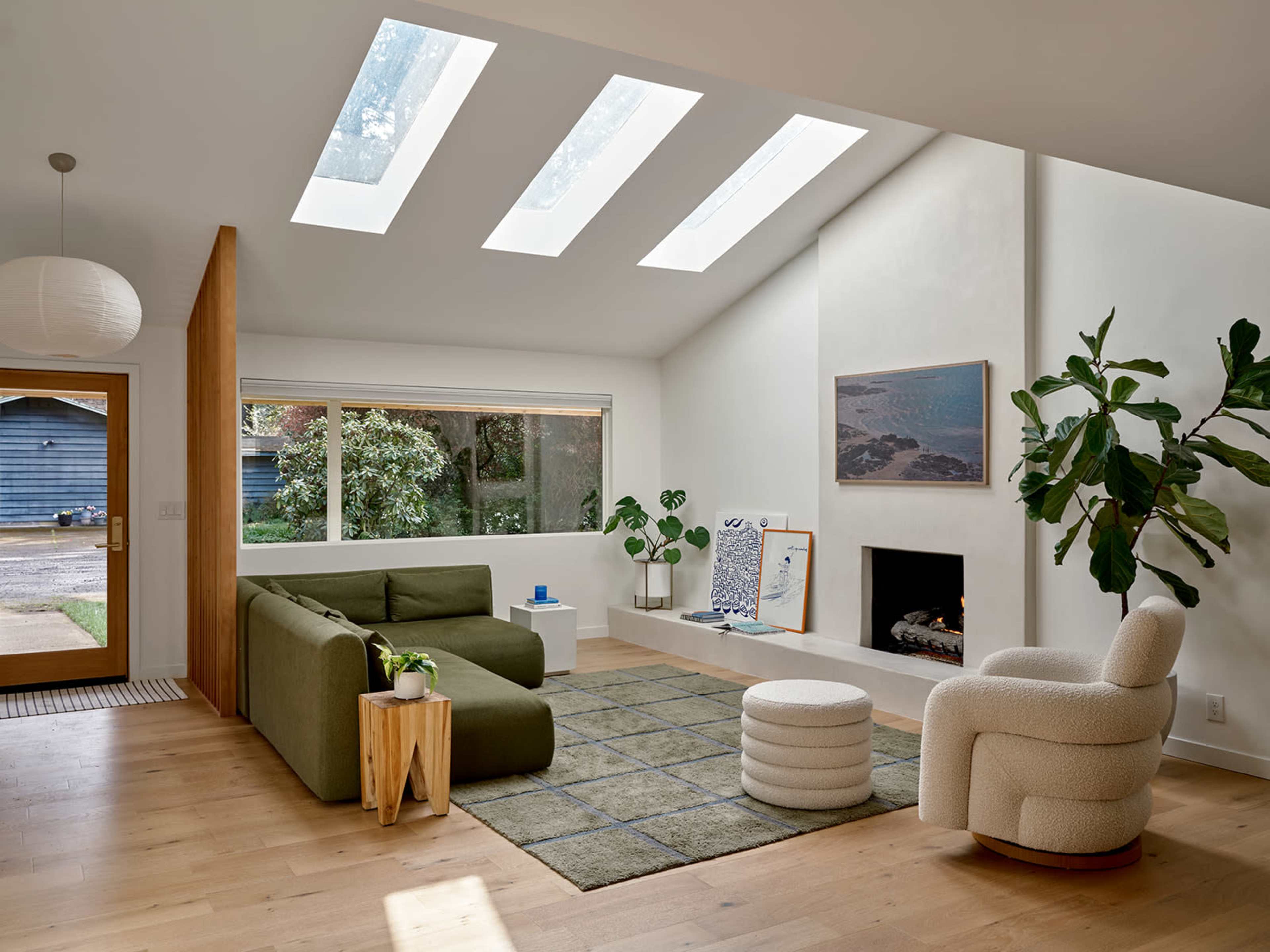 The living room features a large green sectional sofa, a round white pouf, and a tall indoor plant, all set against a backdrop of large windows and a minimalist fireplace.