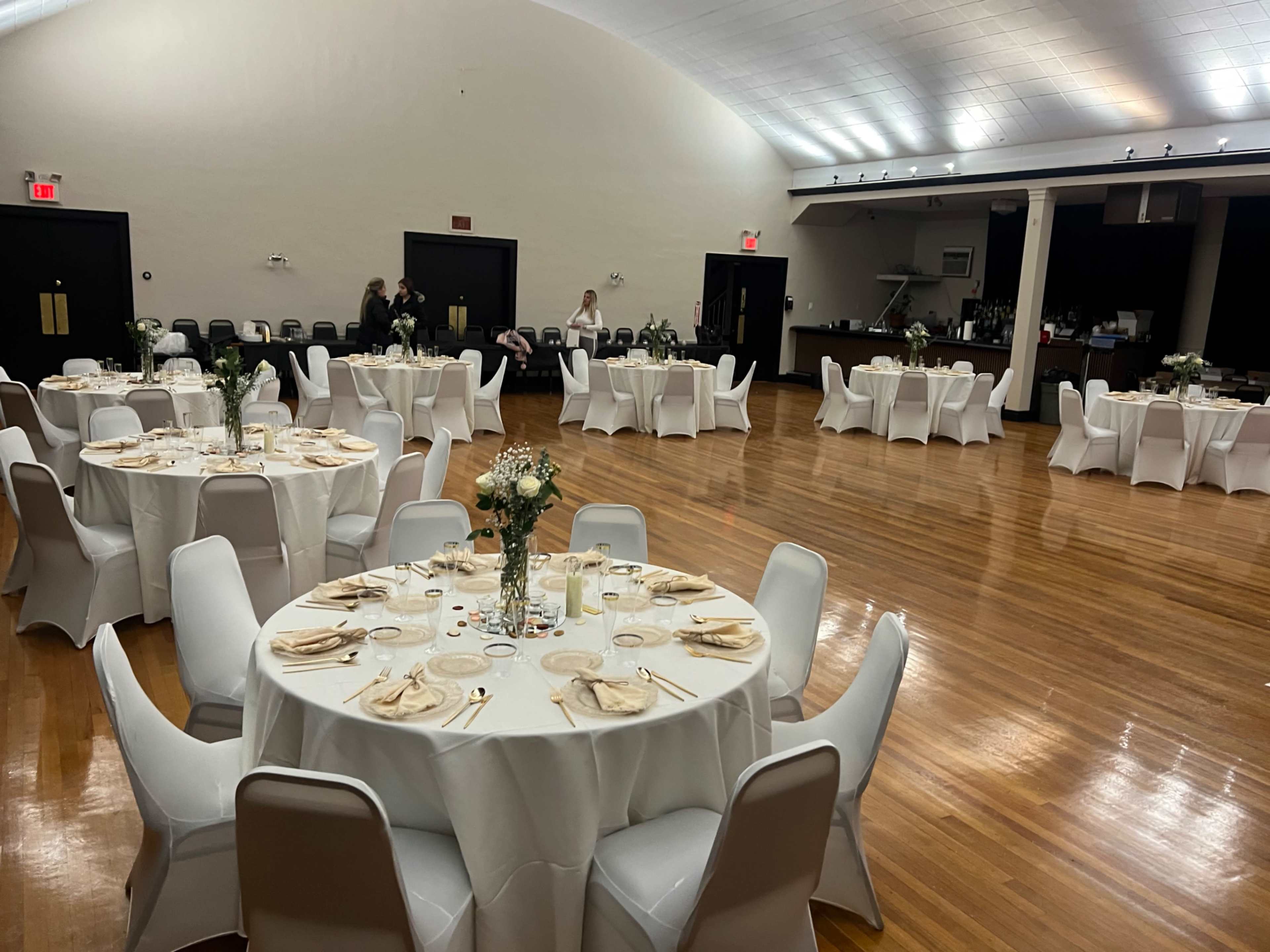 The image shows a spacious hall set up for an event, featuring several round tables covered with white tablecloths, each adorned with floral centerpieces and dinnerware.