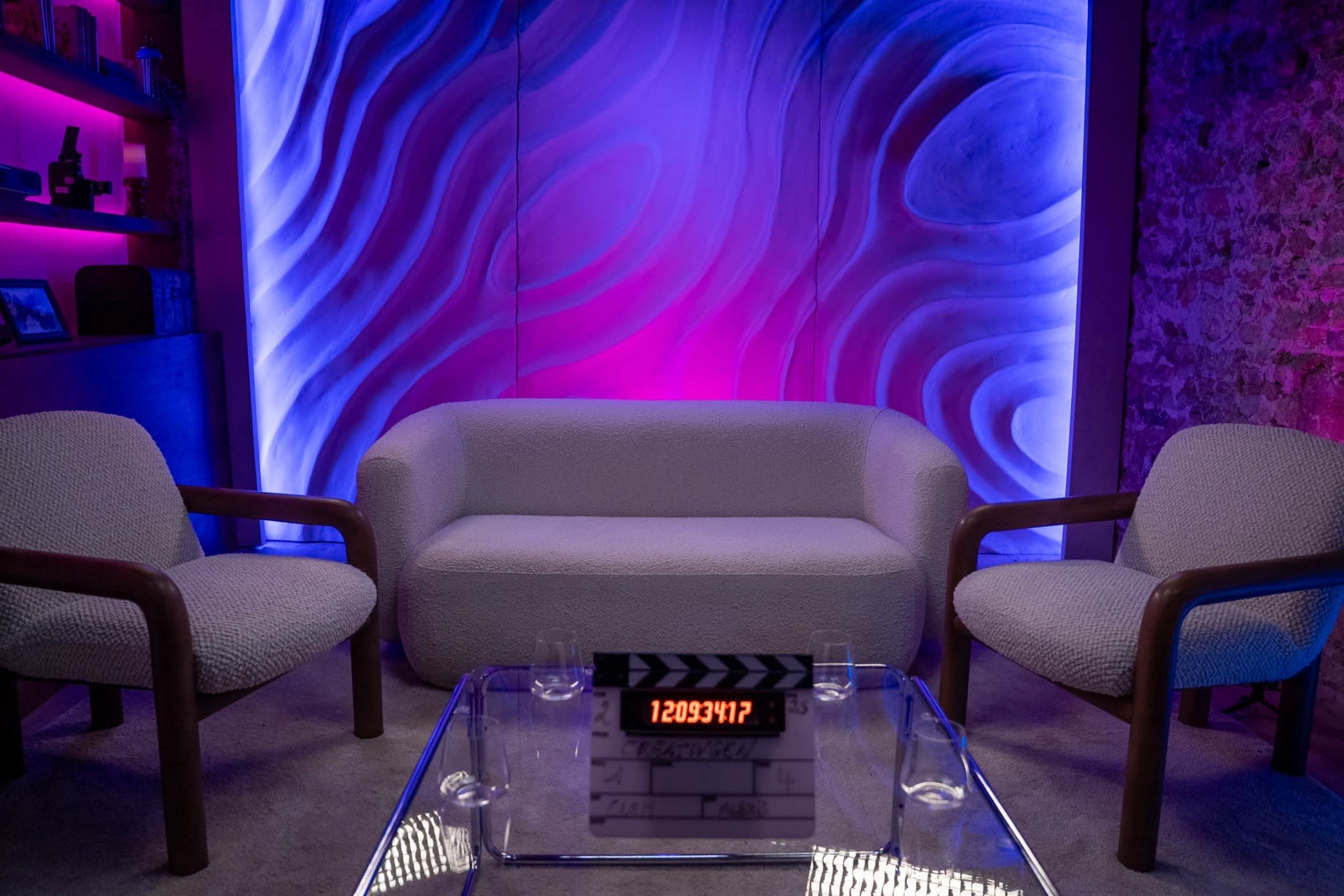 The image shows a modern lounge setting with a light-colored couch, two matching chairs, and a glass table featuring a film clapperboard in front of a colorful, wavy backdrop.