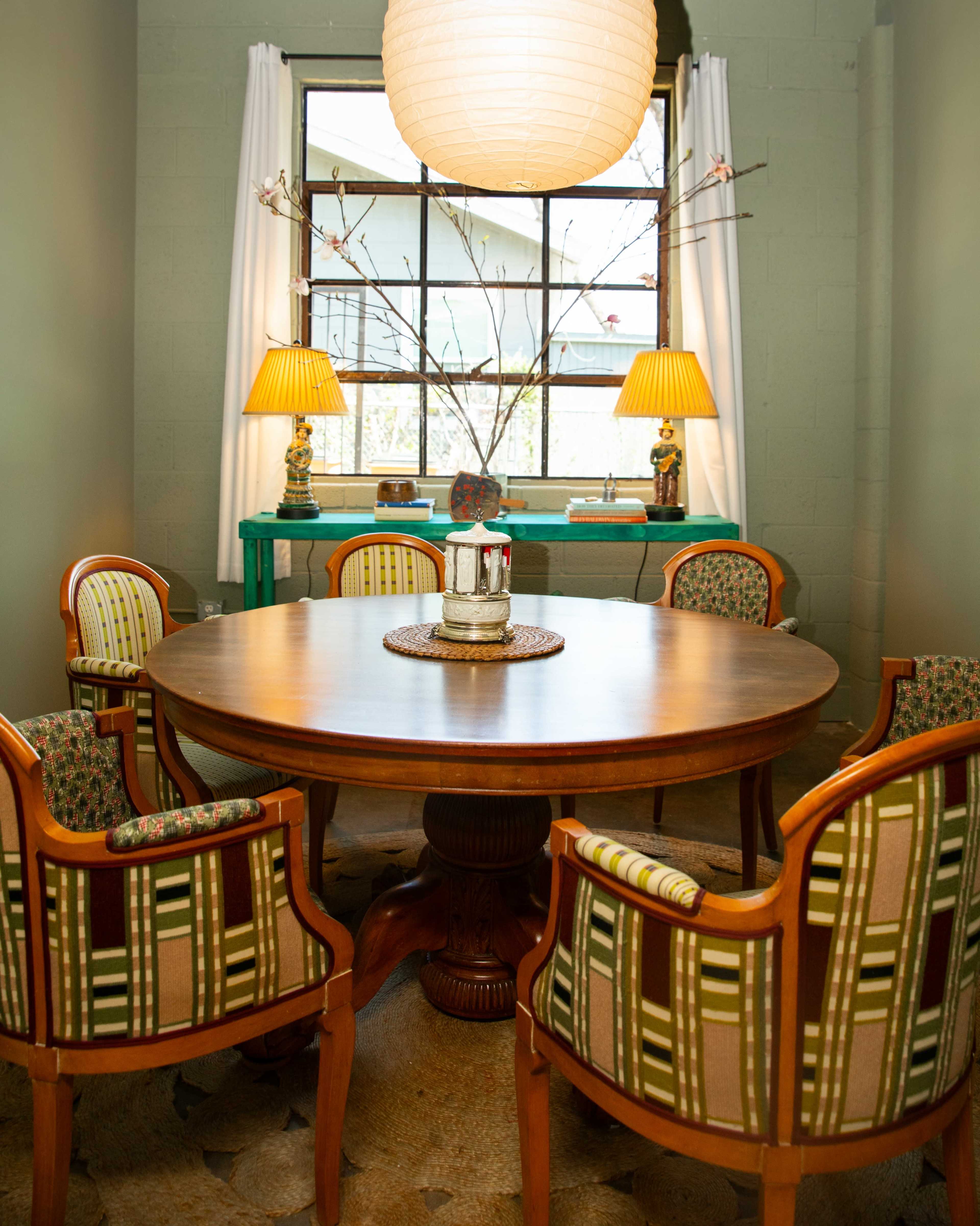 A round wooden dining table surrounded by four upholstered chairs is positioned in a well-lit room featuring a large window and decorative lamps.
