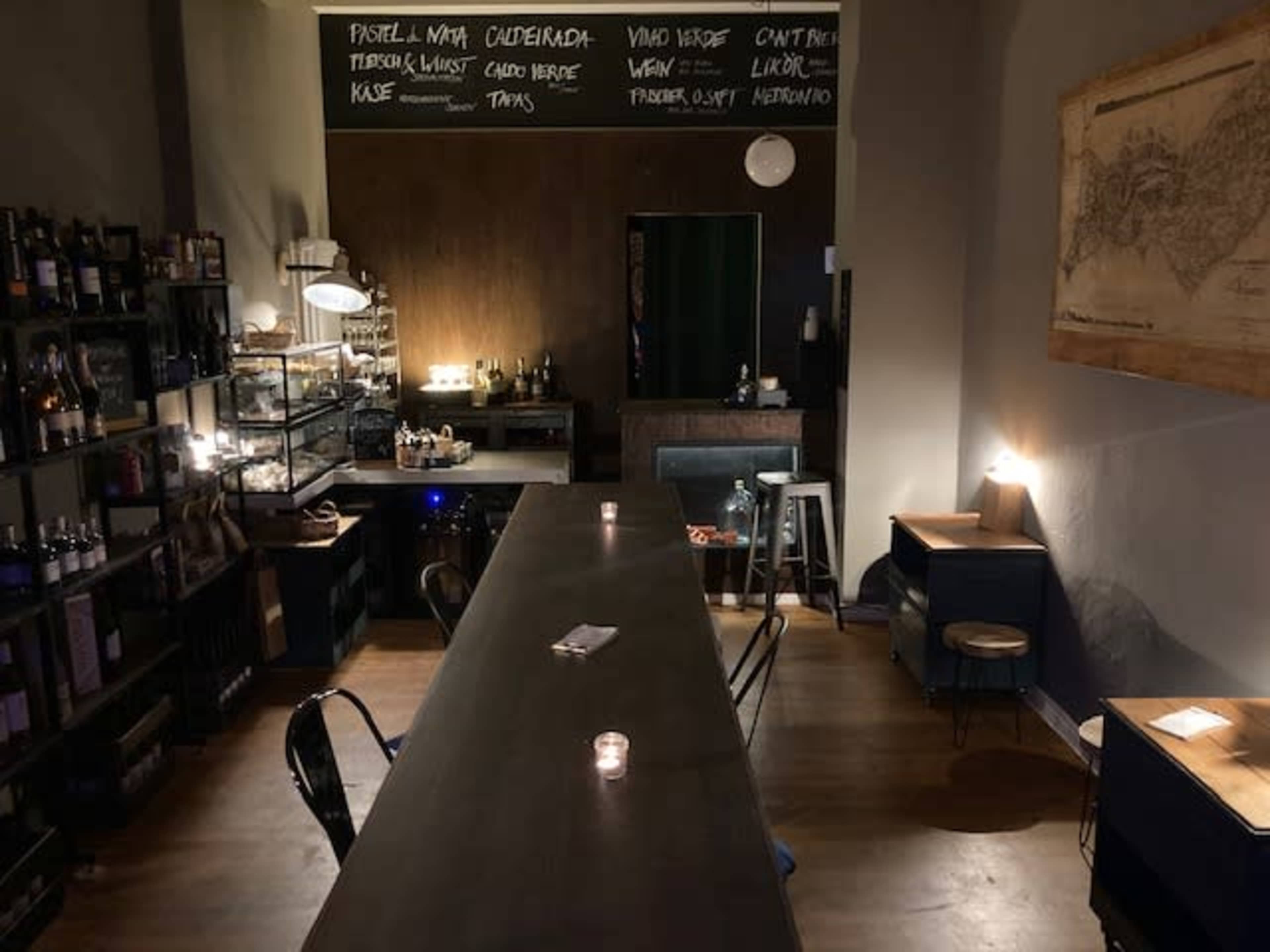 A dimly lit bar area features a long table with candles, shelves lined with bottles, and a menu board on the wall.