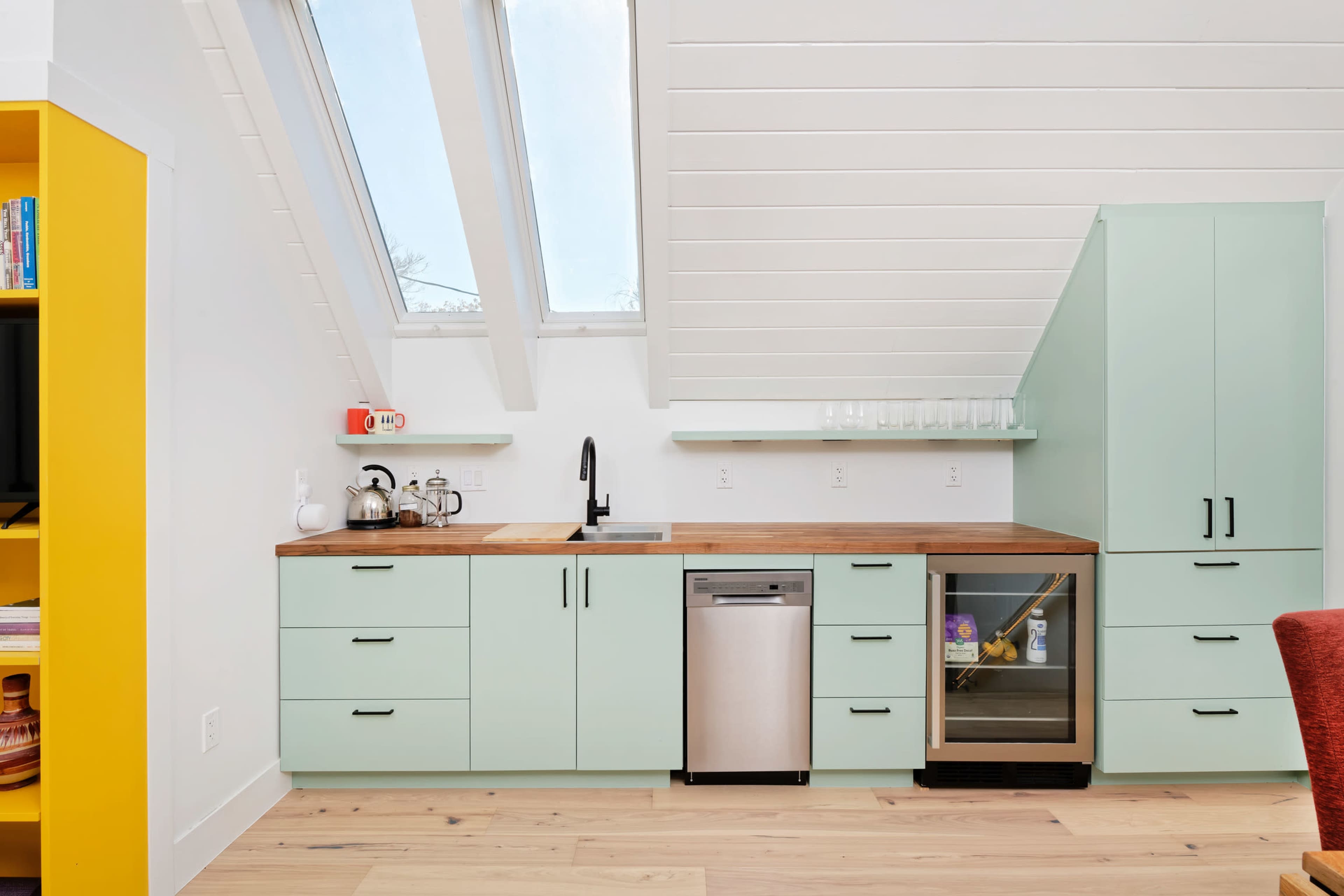 The image shows a modern kitchen with a slanted ceiling, featuring mint-green cabinets, a wooden countertop, and stainless steel appliances.
